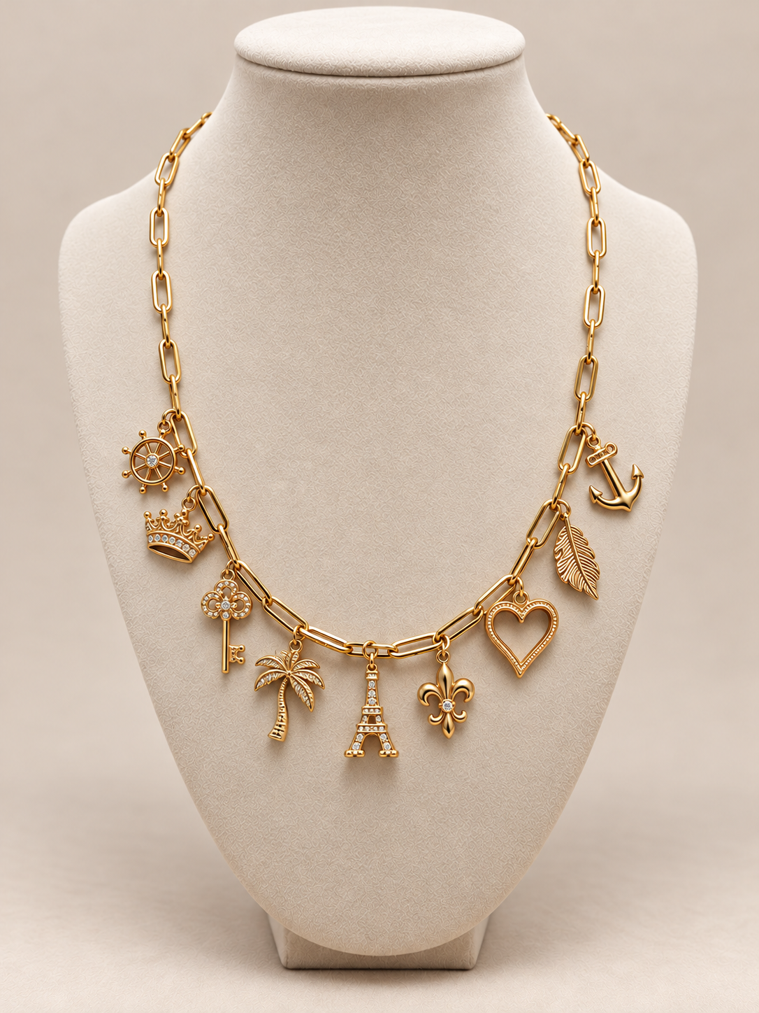 Gold chain necklace with charms including a ship's wheel, crown, flower, key, palm tree, Eiffel Tower, fleur-de-lis, heart, leaf, and anchor on a white display bust.