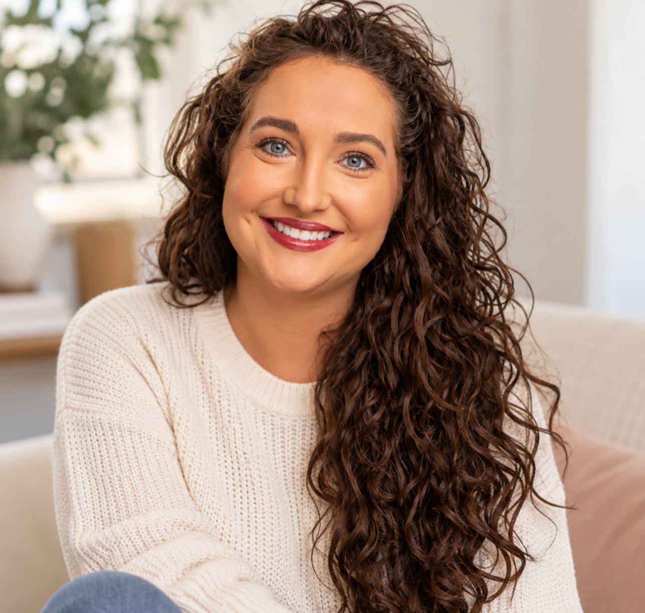 A woman with long curly brown hair and blue eyes smiling, wearing a cream sweater, sitting on a beige couch in a bright room with greenery in the background.