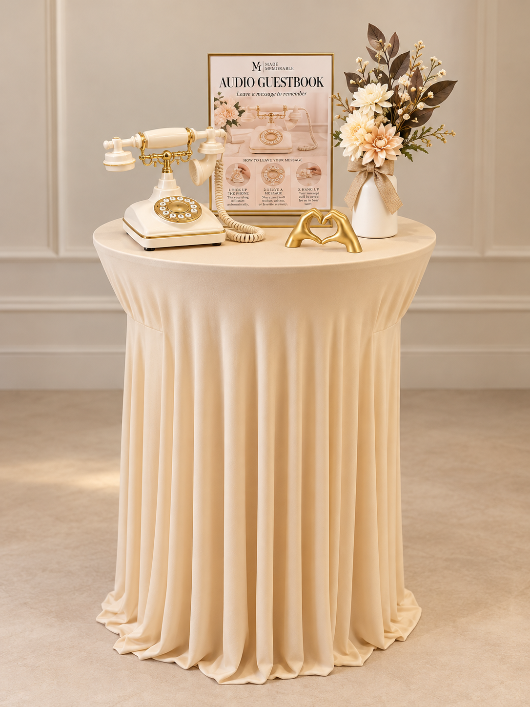 Table with a vintage white and gold rotary phone, a gold hand-shaped decor, a framed informational sign about leaving messages, and a white vase with pink and white flowers and dark green leaves on a beige tablecloth.
