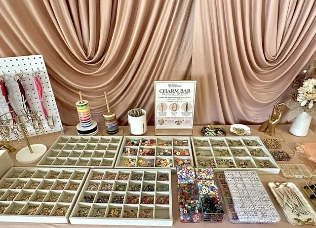 Display of jewelry making supplies, including beads, charms, and tools, on a pink table with a pink curtain background.