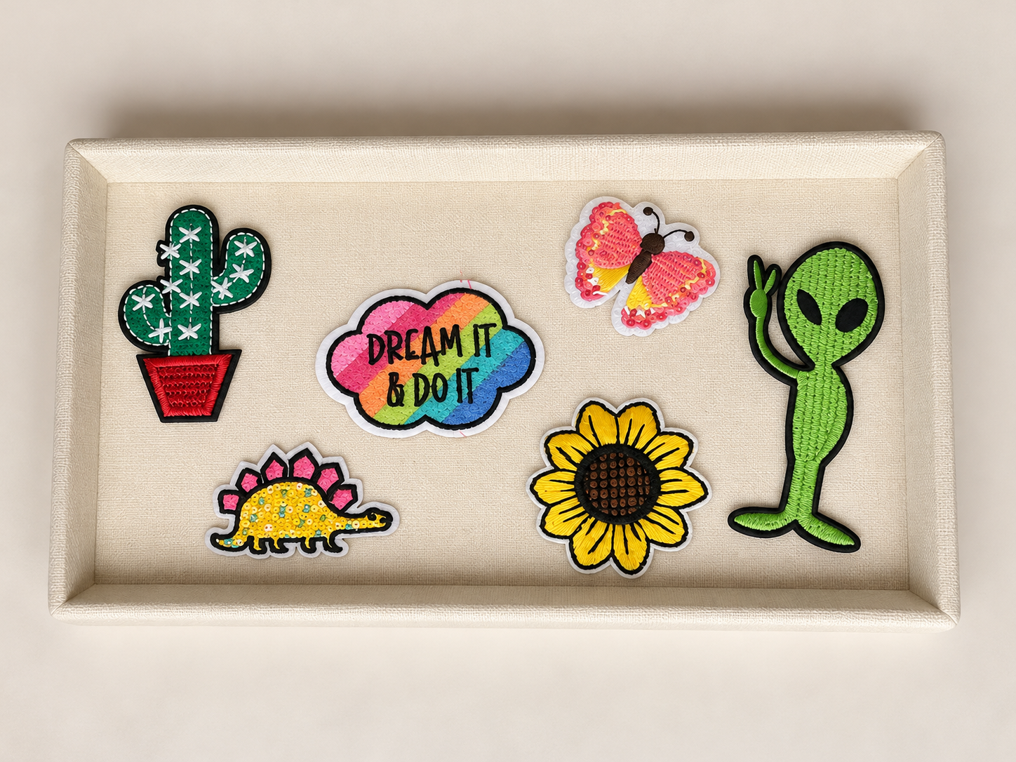 A display of embroidered patches on a beige tray, including a cactus, a rainbow-colored motivational quote, a butterfly, a dinosaur, a sunflower, and a green alien.