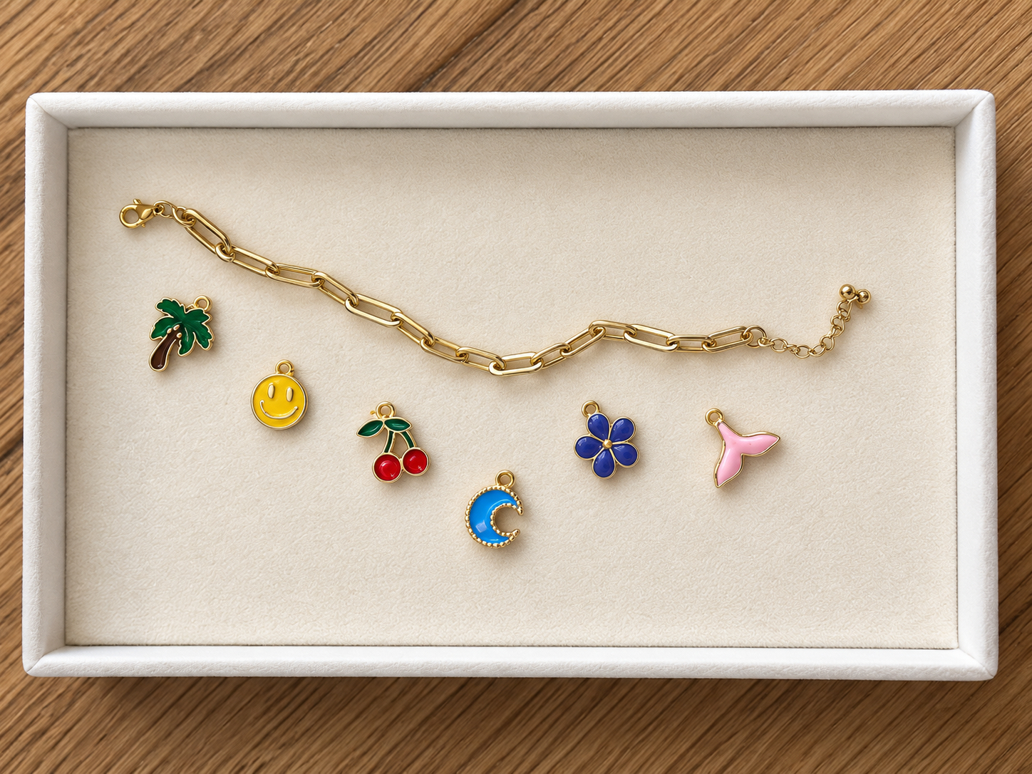 Gold chain bracelet and six colorful enamel charms with nature and whimsical themes inside a white jewelry box on a wooden surface.