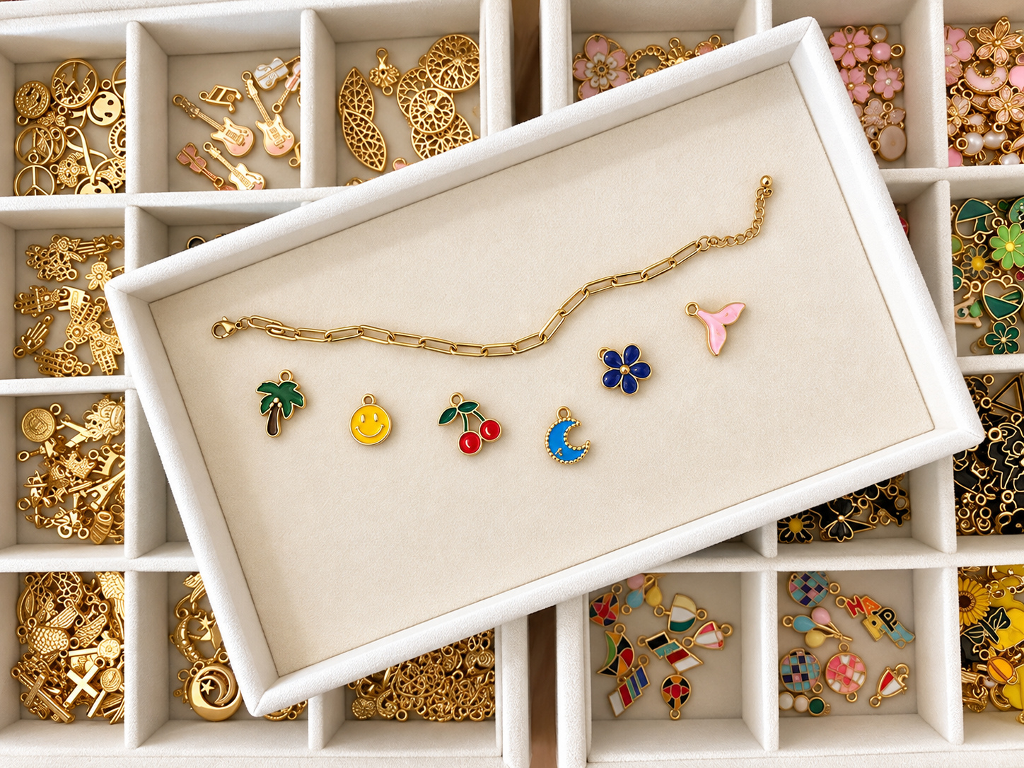A jewelry tray with various colorful charm pendants and a gold chain bracelet on a white display box, surrounded by compartments filled with gold and enamel charm parts and pendants.