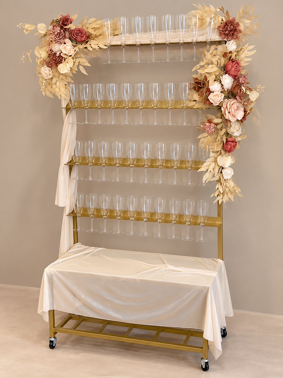 A gold-colored rolling stand decorated with cream, pink, and red paper flowers and beige paper leaves, holding multiple empty champagne flutes arranged on four shelves. A cream fabric drapes over part of the stand.