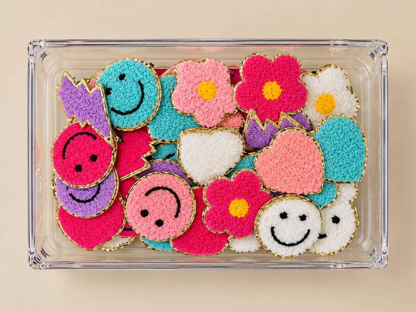 A clear plastic container filled with colorful, embroidered cookies in the shapes of smiley faces, hearts, and flowers, with gold trim around the edges.