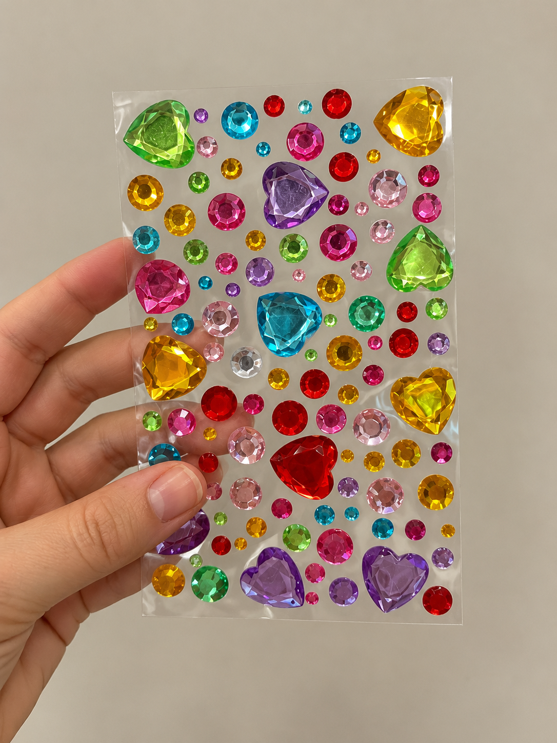 A hand holding a clear sheet of colorful heart and round-shaped adhesive rhinestones against a plain background.