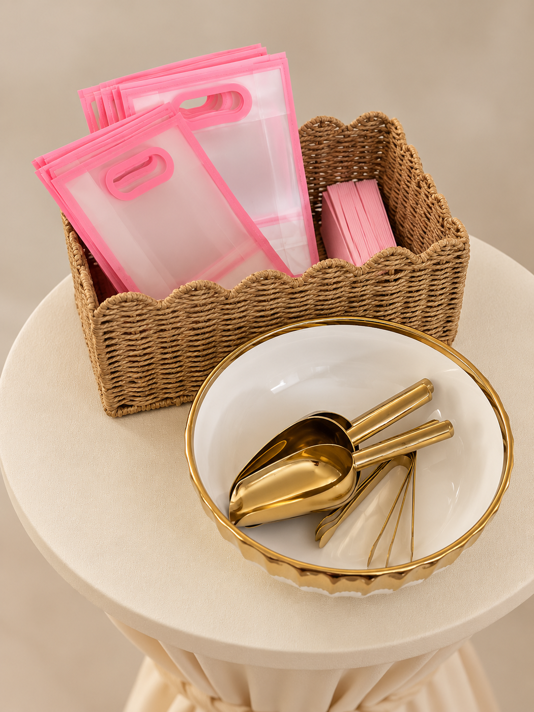 A wicker basket with pink plastic sandwich bags and pink paper napkins sits on a small, round beige table. Next to it is a white bowl with a gold rim containing gold-colored scoops, tongs, and serving utensils.