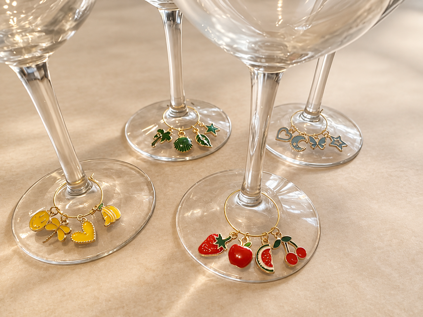 Four empty wine glasses on a beige surface, each decorated at the base with colorful charms, including fruits, plants, and abstract shapes.