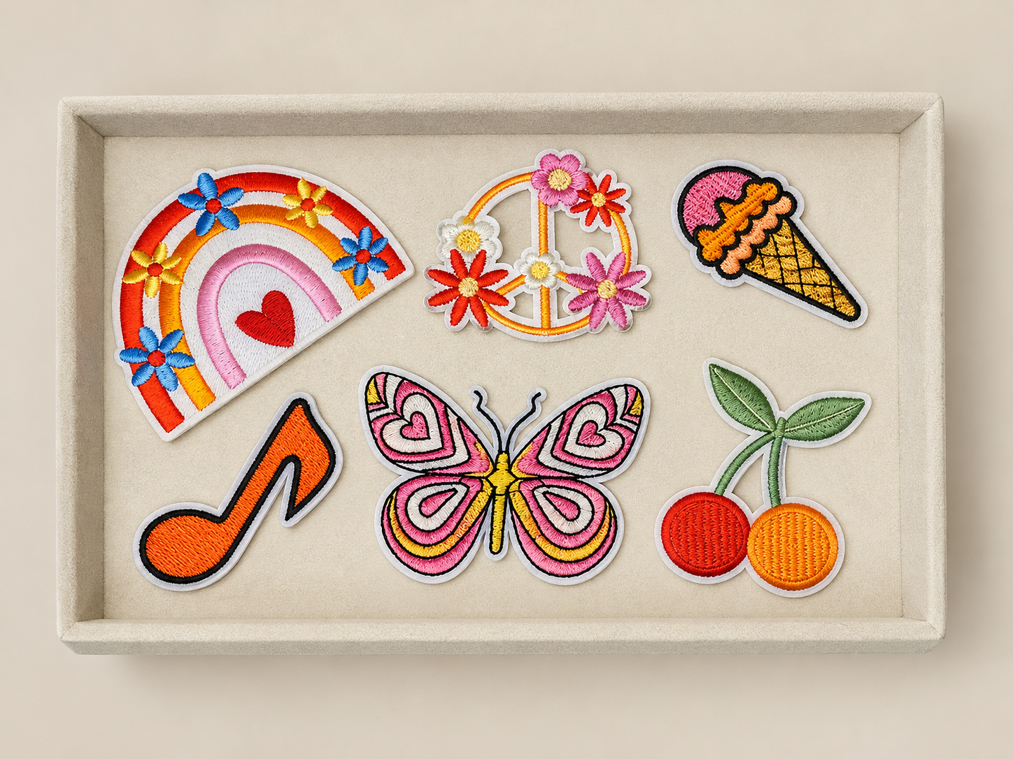 Set of colorful embroidered patches including a rainbow with a heart, peace sign with flowers, ice cream cone, musical note, butterfly, and cherries, arranged in a display box.