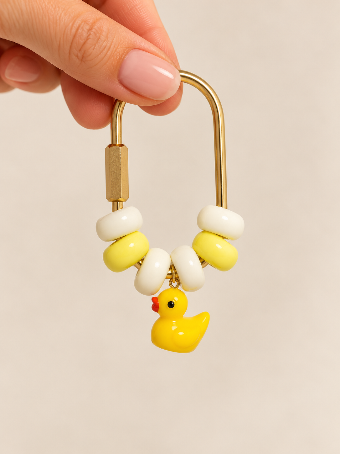 A hand holding a gold-colored safety pin decorated with white and yellow beads and a small yellow rubber duck charm.