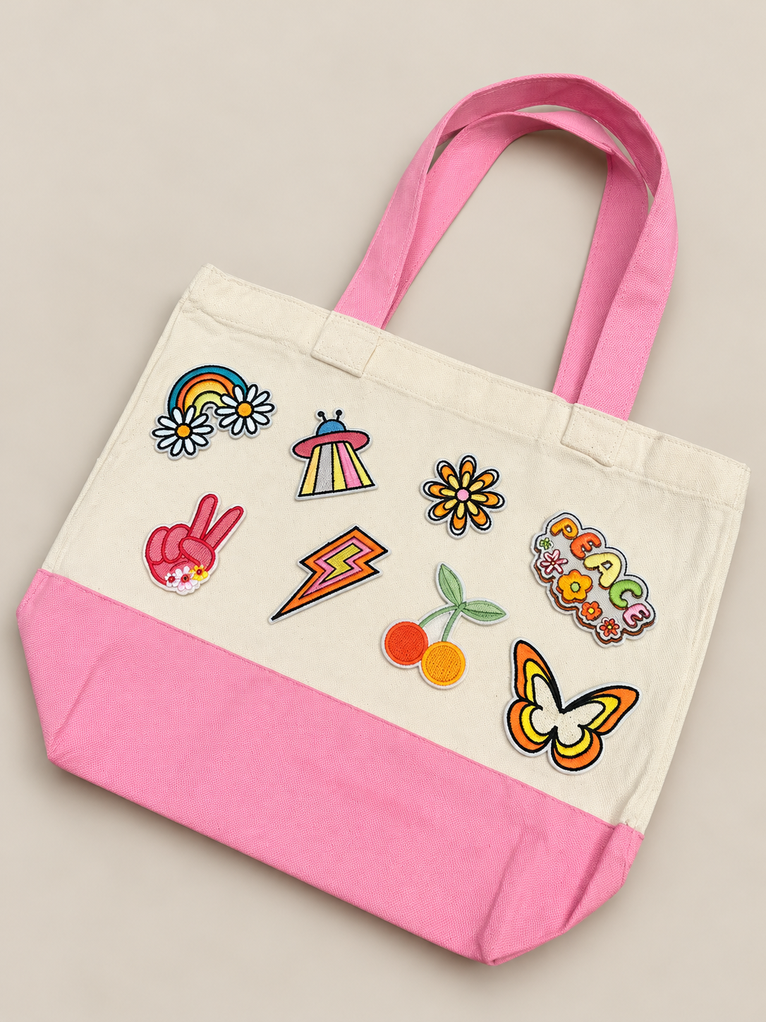 Canvas tote bag with pink handles and bottom, decorated with colorful patches including rainbow, UFO, flowers, peace sign, lightning bolt, cherries, butterfly, and the word 'PEACE'.