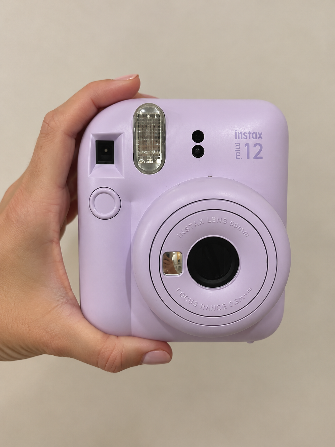 A person holding a lavender Fujifilm Instax Mini 12 instant camera with a 60mm lens, flash, and viewfinder, against a plain background.