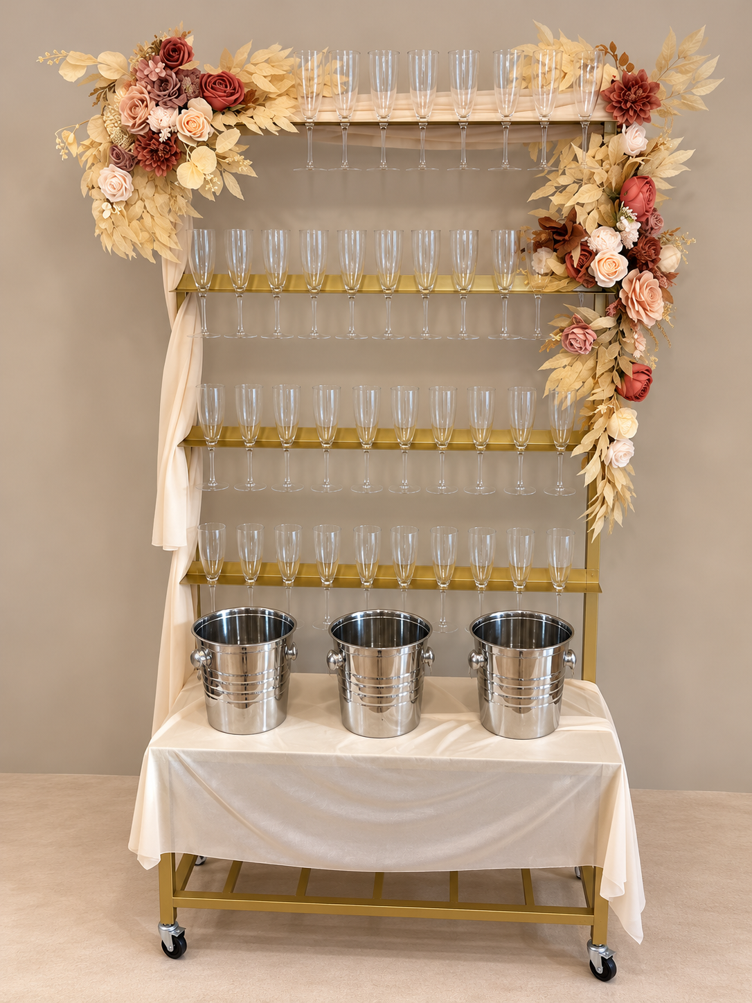 Champagne flute display with floral decorations on a gold-colored stand, featuring five empty champagne glasses on top, five in the middle, and four at the bottom, with three metal buckets on a table draped with a white cloth.