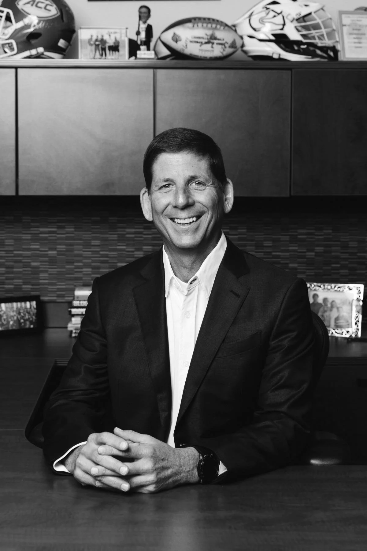 A black and white photo of a smiling man in a business suit sitting at a desk with hands clasped. Behind him is a cabinet with football helmets, framed photo, and various objects.