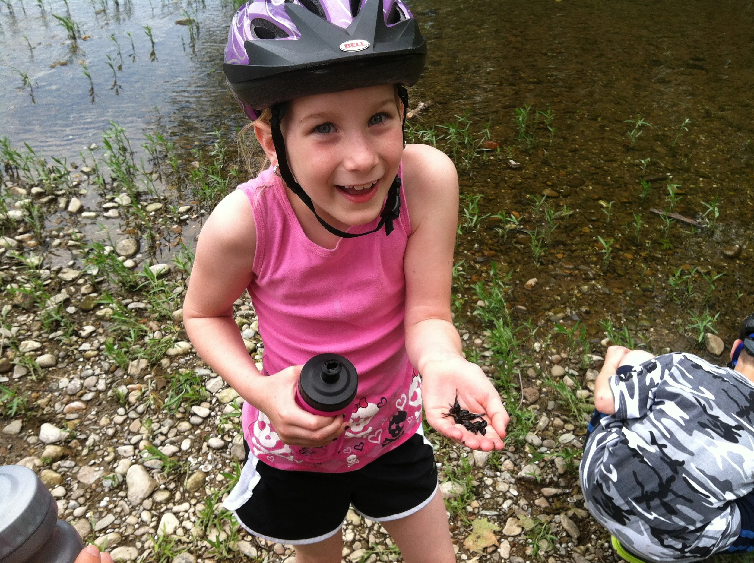 A young girl with blue eyes and freckles wearing a purple bike helmet and pink sleeveless top, holding a bottle and some insects, kneeling by a riverbank with small green plants and rocks.