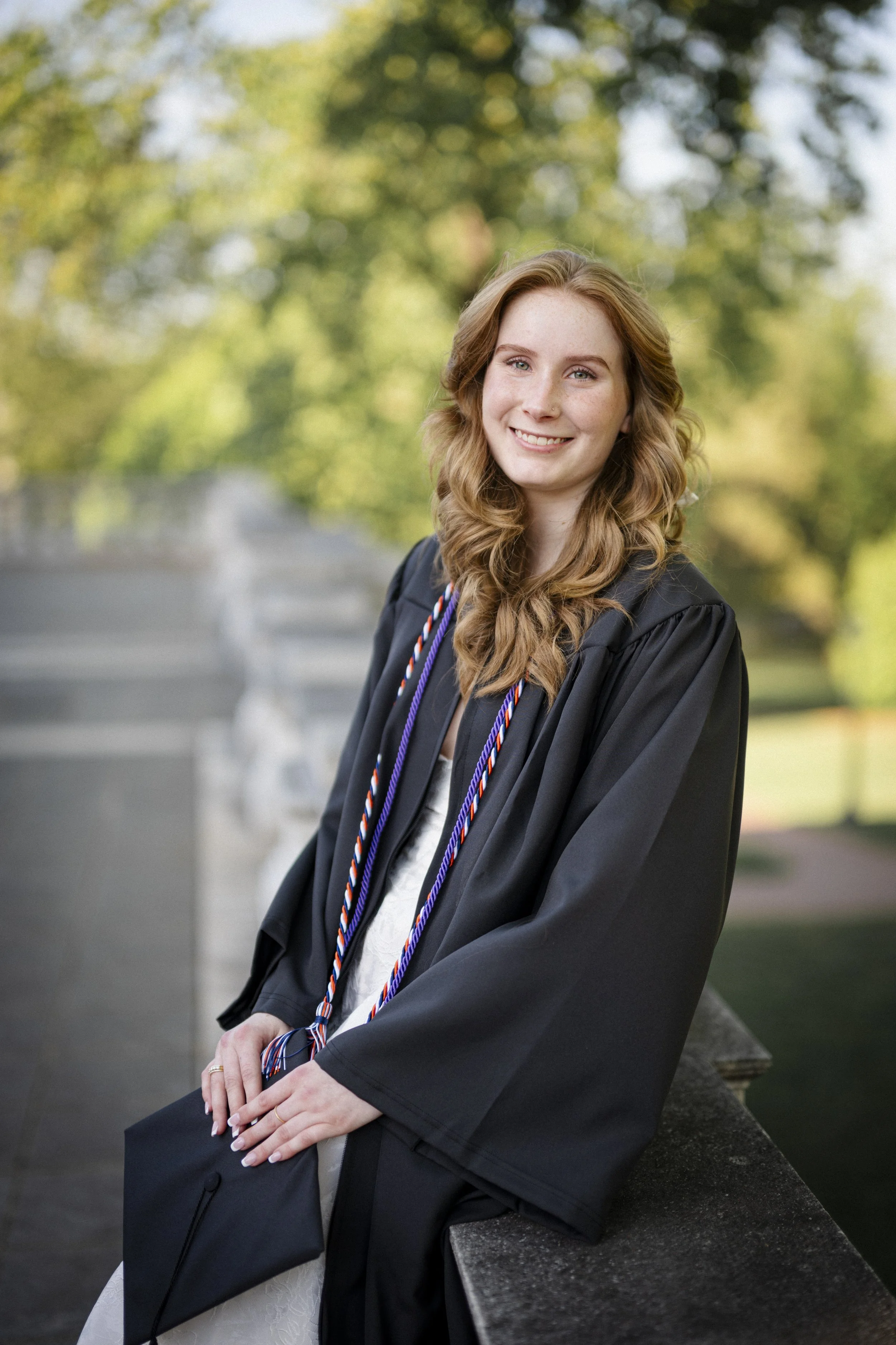 A young woman in a black graduation gown and cap, holding her cap in her hands, sitting on a stone ledge in a park with blurred green trees in the background, smiling at the camera.
