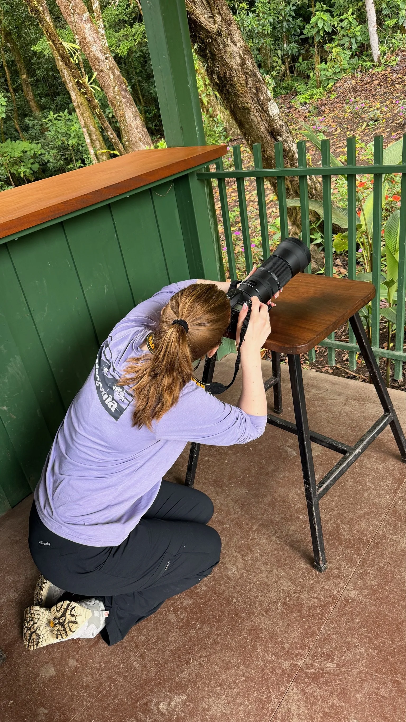 A woman crouching with her head against a green wooden fence, looking through a camera with a long lens, set on a table outdoors in a lush, forested area.