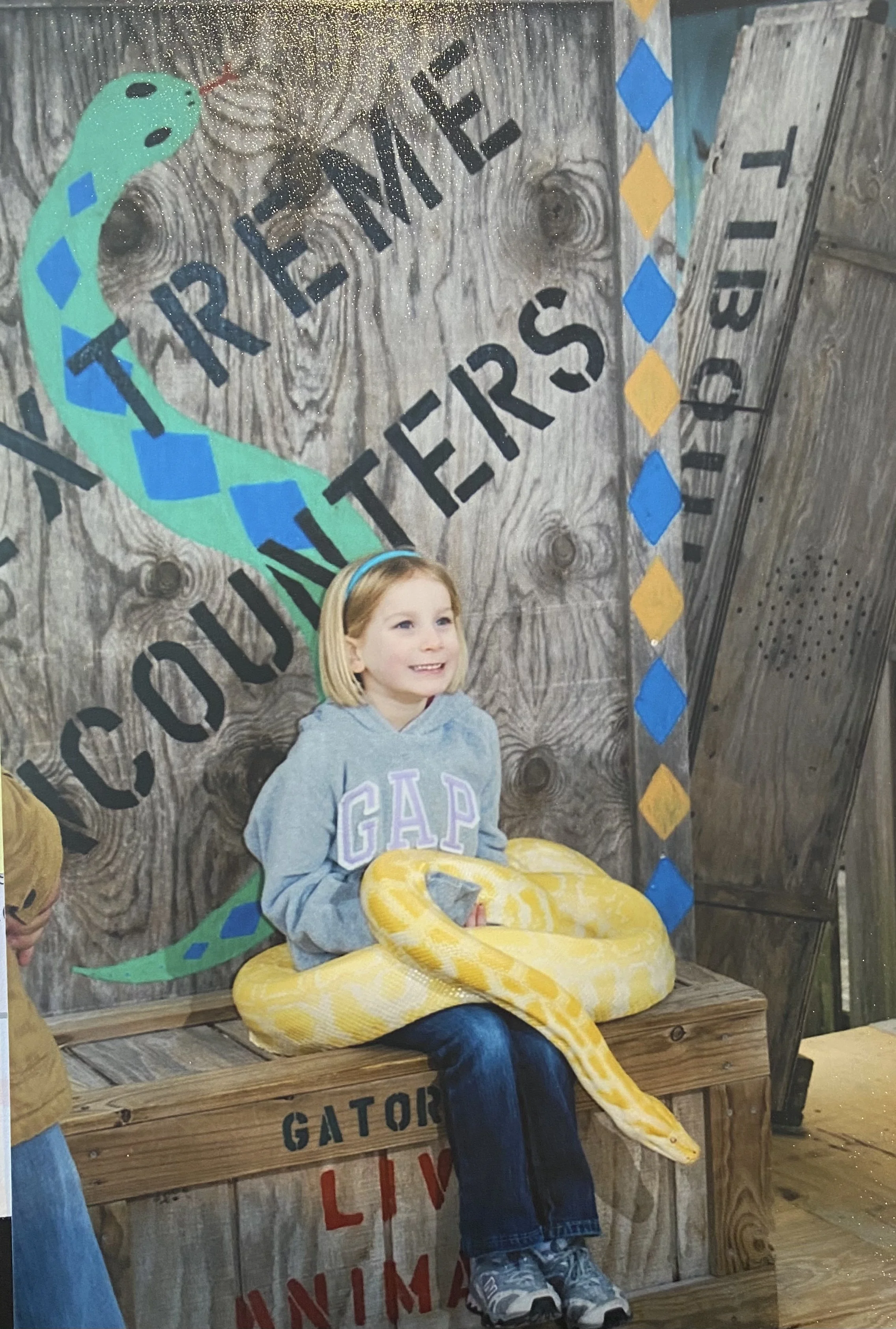 A young girl smiling while sitting on a wooden platform, holding a large yellow and white albino python. Behind her is a wooden wall with painted signs including 'TRIPLE', 'GATOR', and 'LIVIN'.