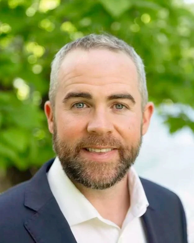 A headshot of a man with short gray hair, beard, and blue eyes, wearing a dark suit and white shirt, outdoors with green trees in the background.