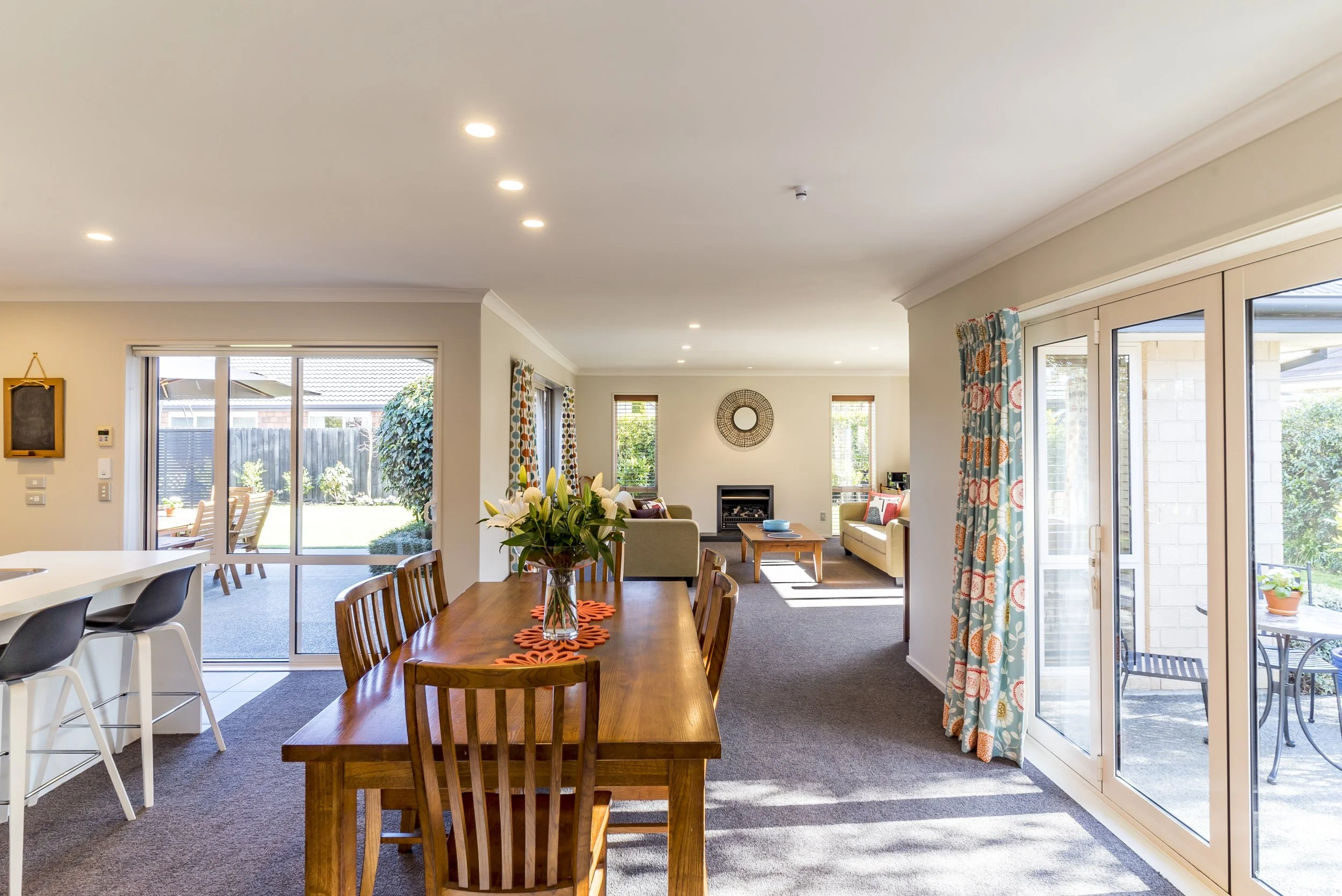 A spacious living room with a wooden dining table, six chairs, a bouquet of white lilies, a tv on a stand, multiple windows with curtains, and glass sliding doors leading outside to a backyard with patio furniture and green plants.