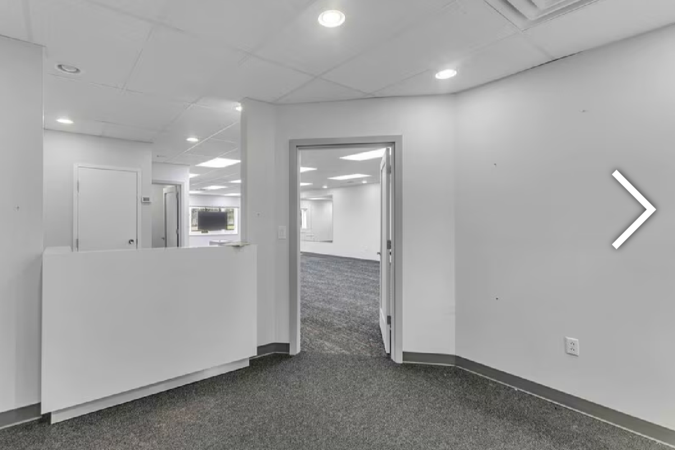 Empty office with white walls, gray carpet, ceiling lights, and a partial glass door leading to another room.