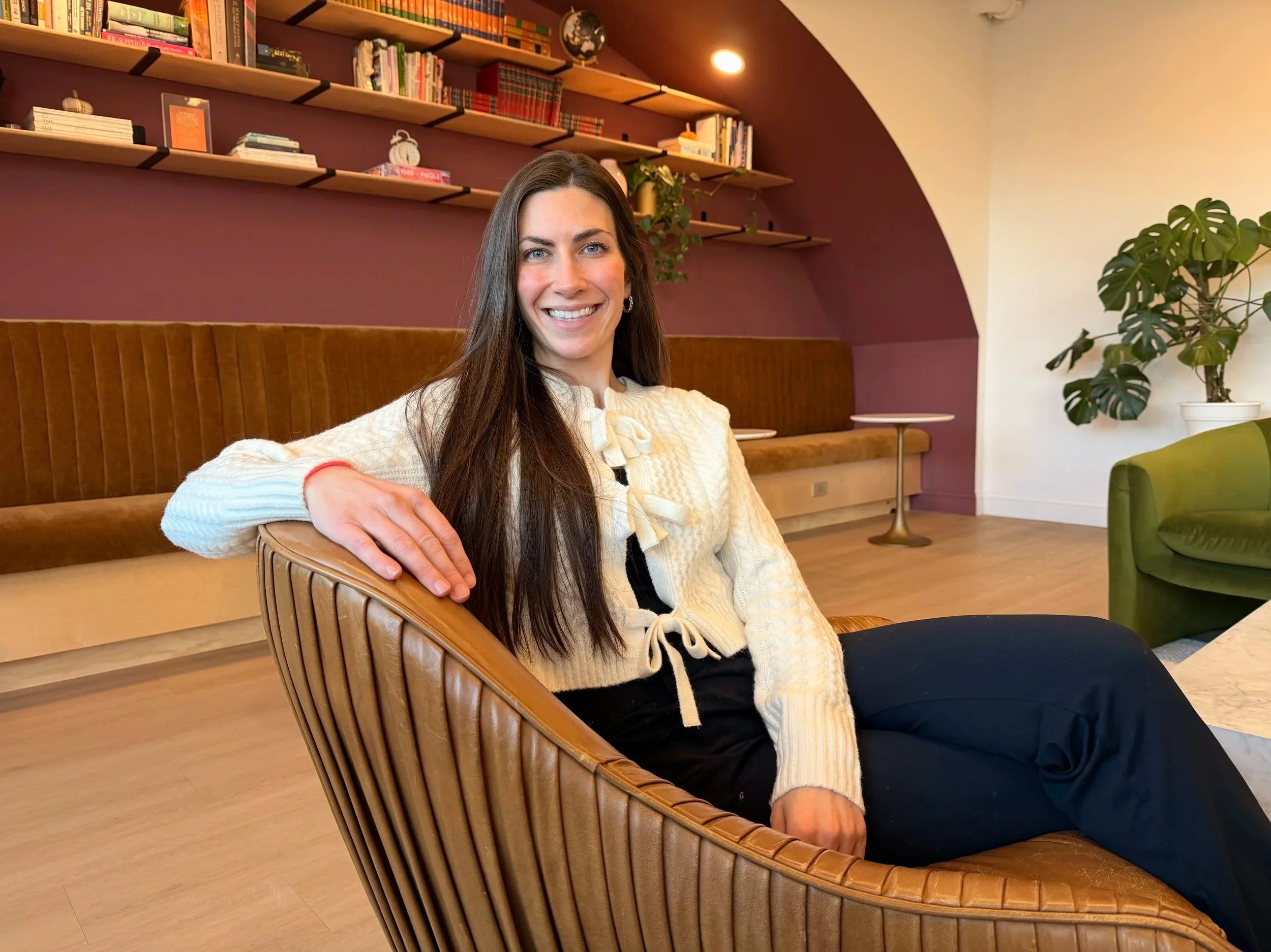 A woman with long brown hair and blue eyes smiling and sitting on a wooden chair in a cozy room with bookshelves, plants, and modern decor.