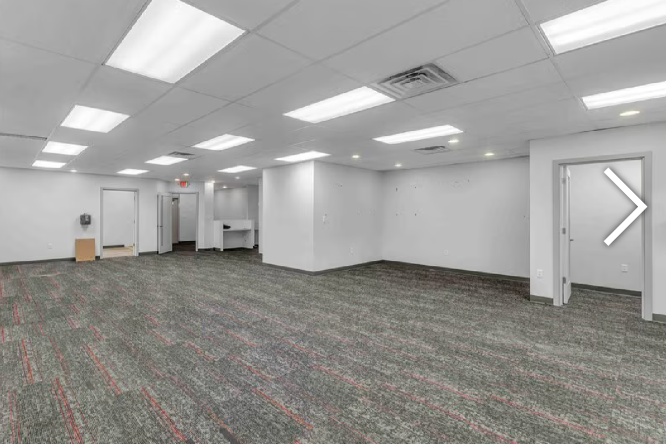 Empty office space with white walls, gray patterned carpet, and fluorescent ceiling lights