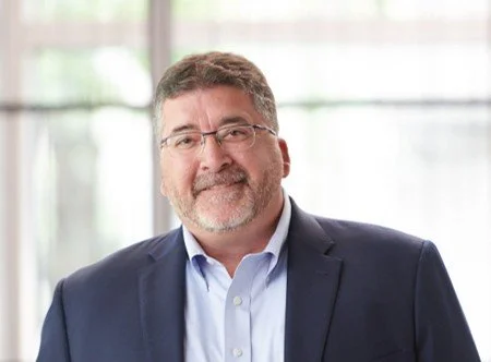 Portrait of a middle-aged man with glasses and a beard, wearing a navy blazer and light blue shirt, standing indoors with windows in the background.