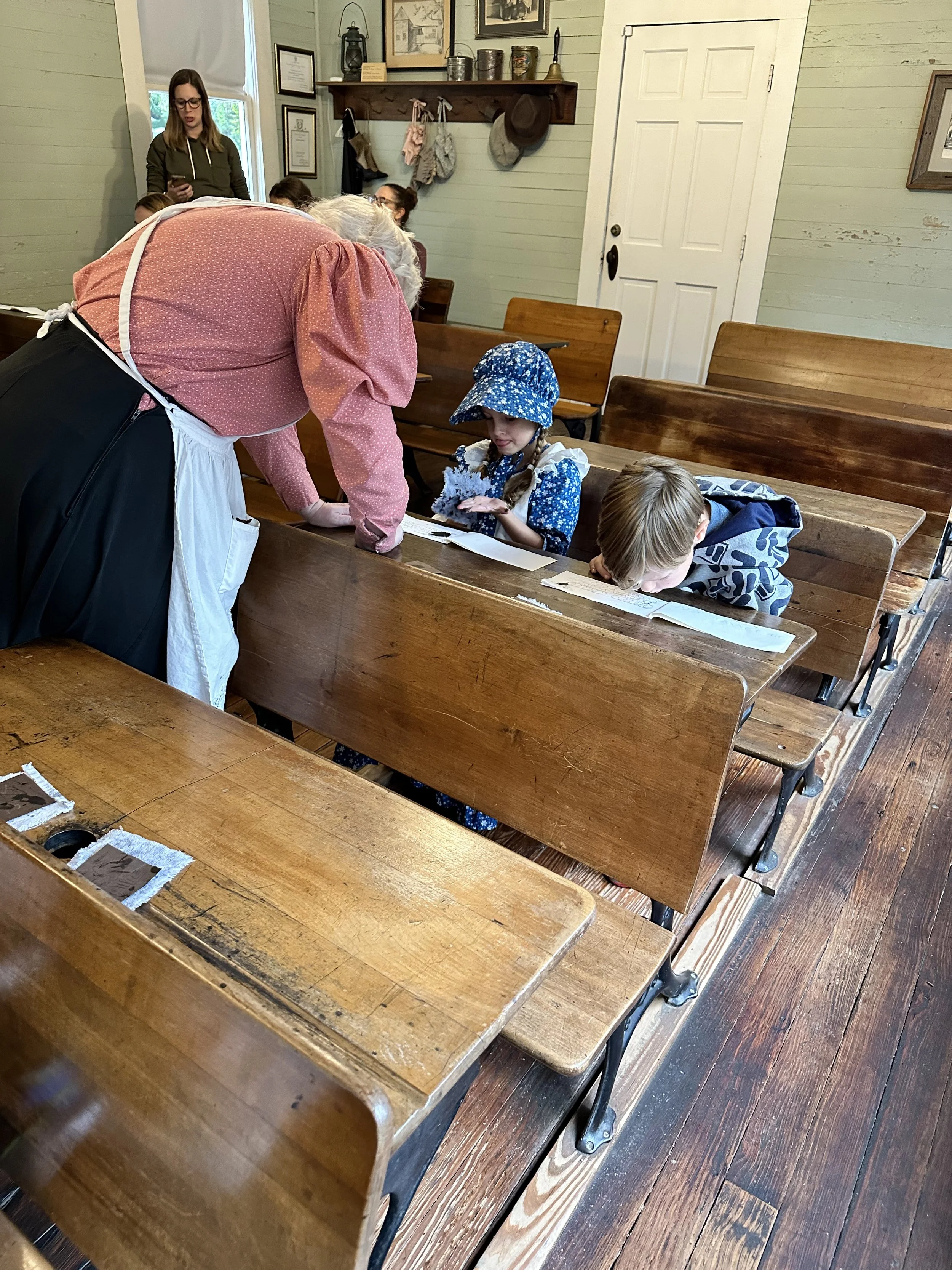 An elderly woman with gray hair leaning over a wooden pew, writing on paper, with children sitting at the pew and a woman standing in the background inside a rustic room with wooden walls, framed pictures, and a shelf with hats and jars.