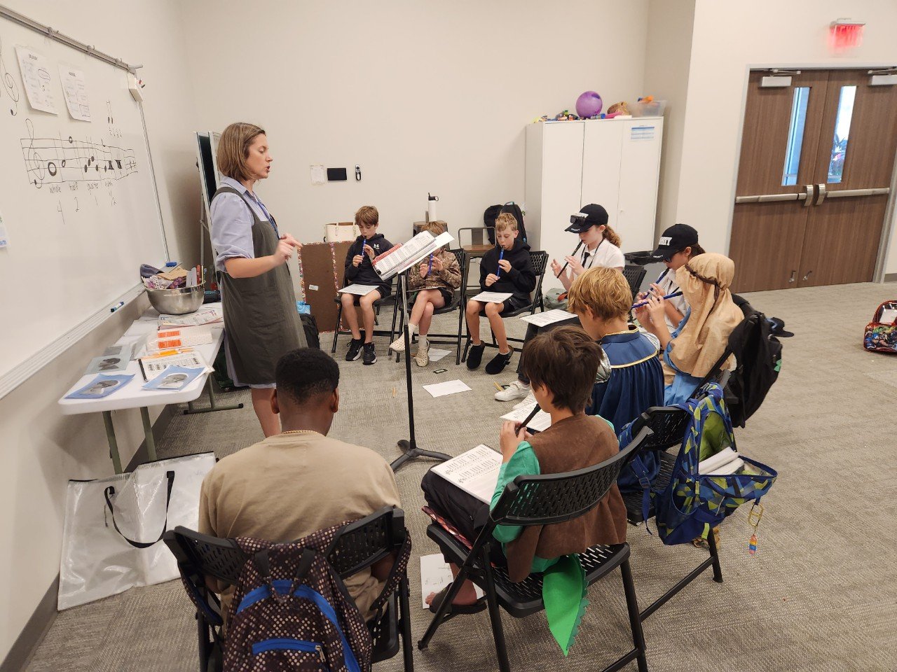 A music teacher giving a lesson to a group of children sitting in a semi-circle, playing recorders in a classroom setting.