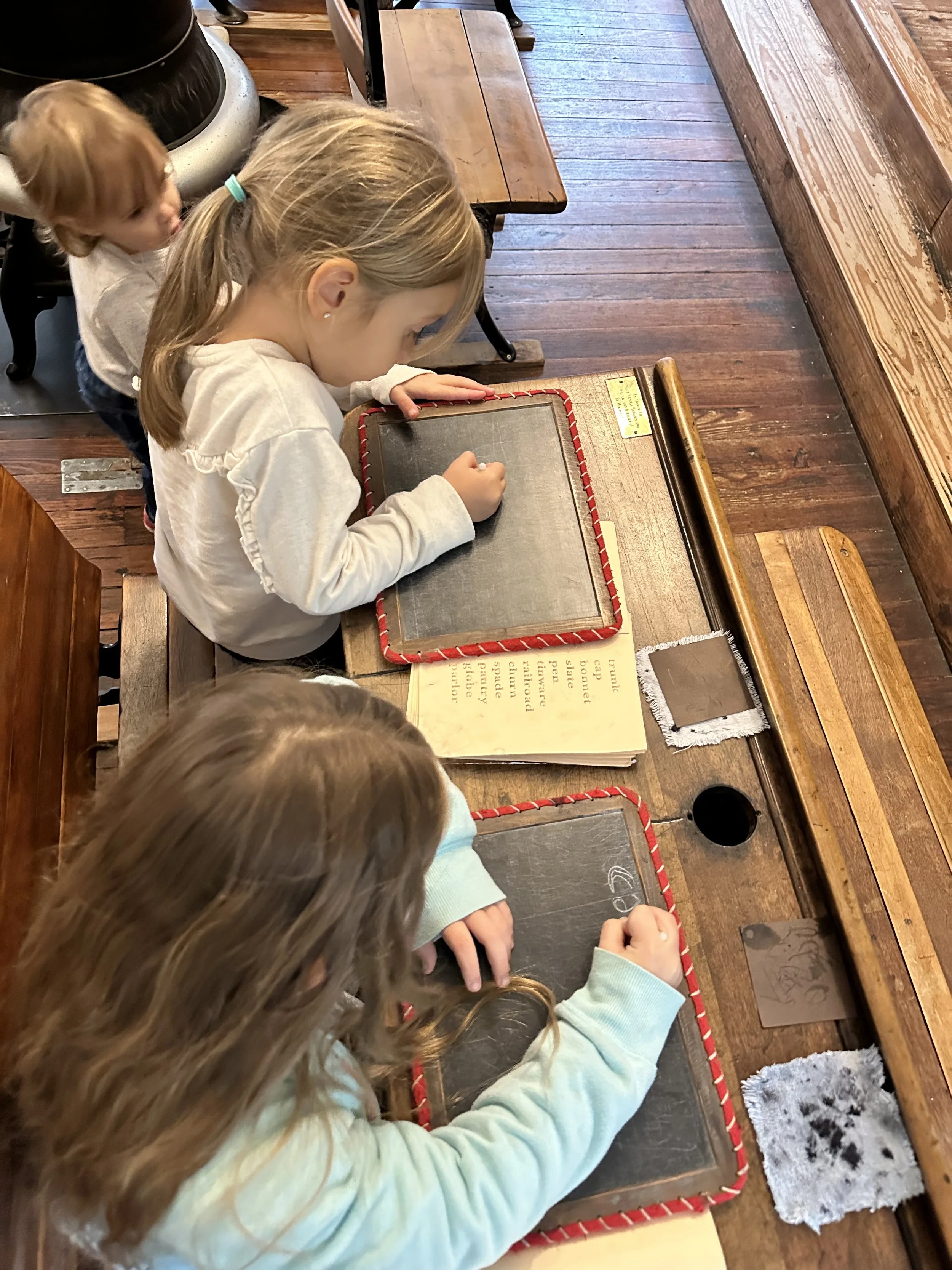 Children sitting at a wooden table in a classroom, drawing on small chalkboards with chalk.
