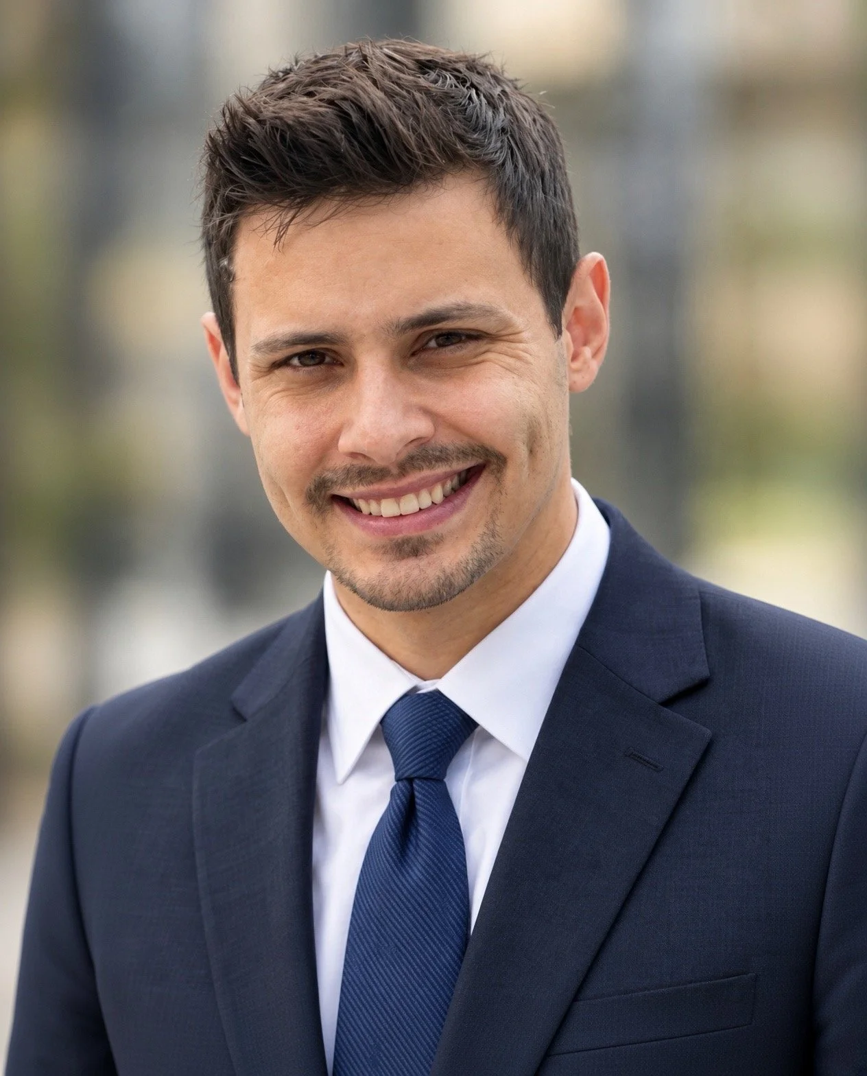 A man in a navy blue suit with a blue tie, smiling outdoors with a blurred background.