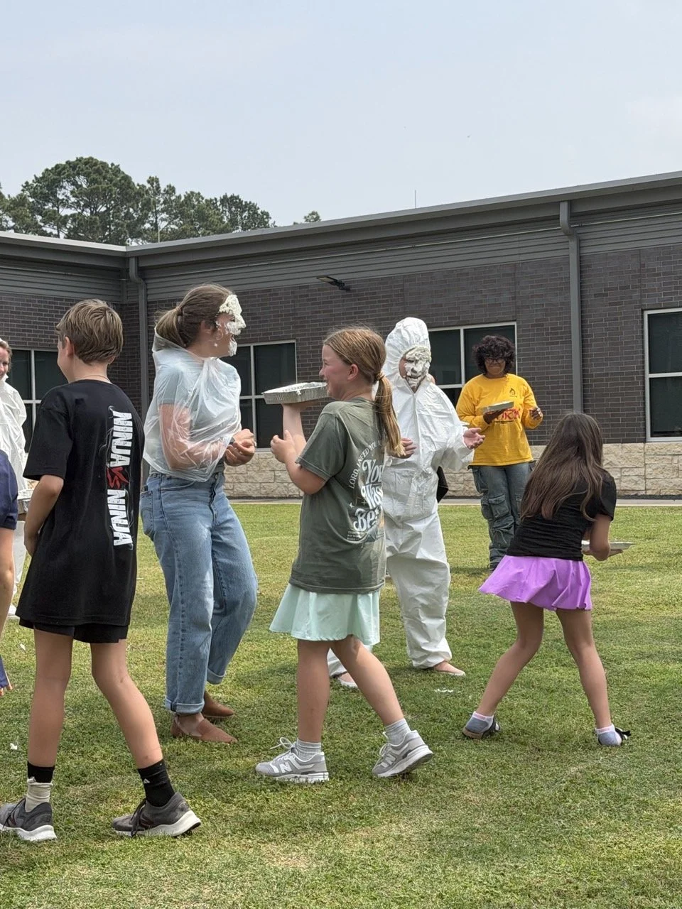 Children and adults engrossed in a pie-throwing game outdoors, with children preparing to throw pies at adults with whipped cream on their faces, on a grassy area near a brick building.