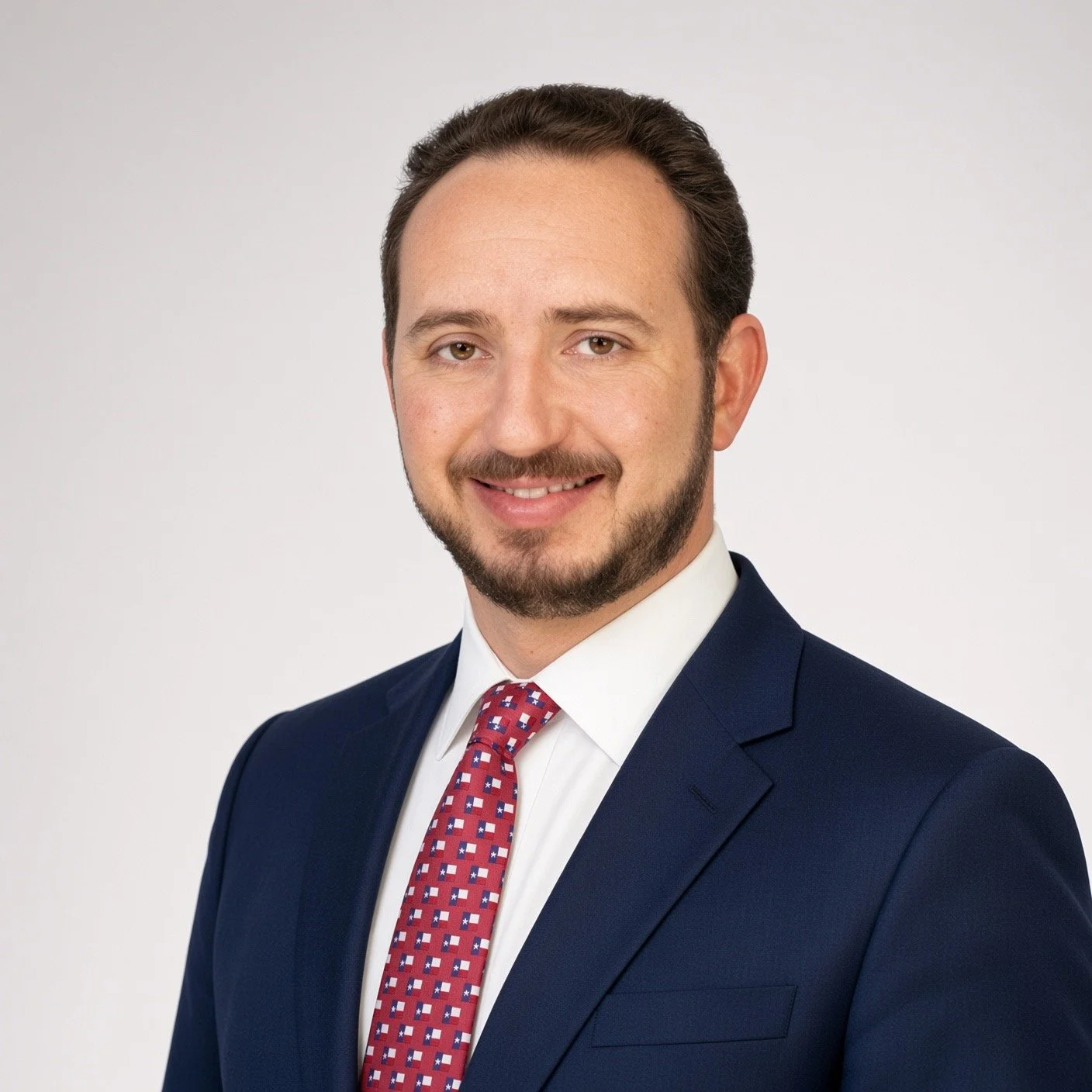 Headshot of a man with brown hair and a beard, dressed in a dark blue suit, white shirt, and red patterned tie, smiling against a plain white background.