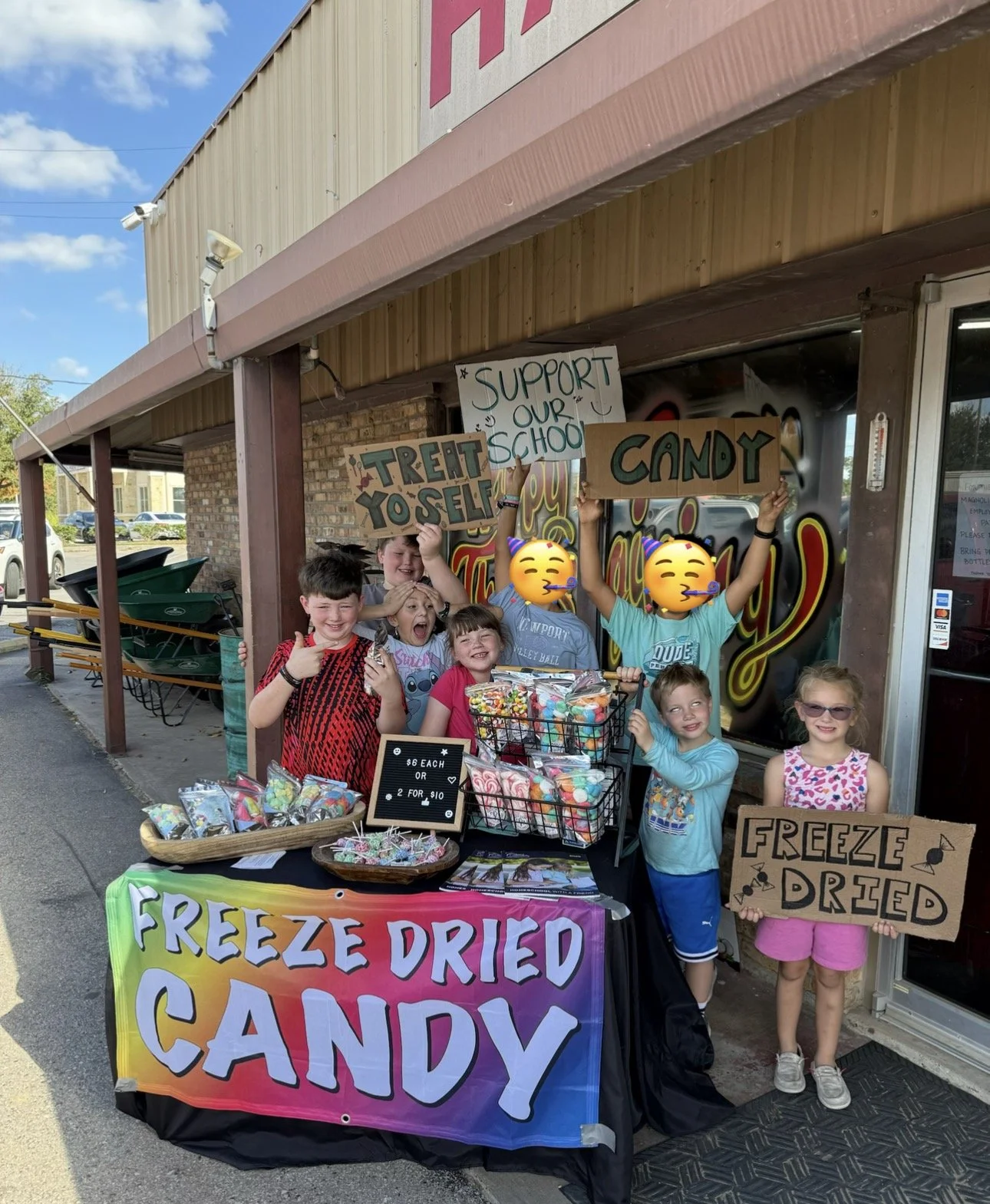 Children selling freeze-dried candy outside a store with signs that read 'Support Our School,' 'Candy,' and 'Freeze Dried.' The children are smiling and holding signs, with colorful candy displays on the table in front. The store has a brick and metal exterior, and a rainbow-colored banner that says 'Freeze Dried Candy.'