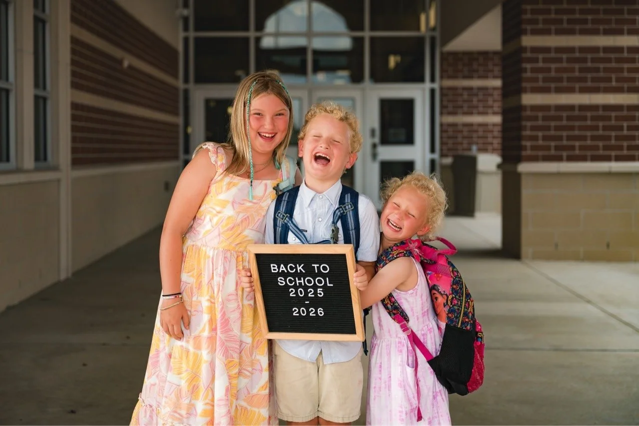 Four children standing outside a school building, smiling and hugging. The boy in the middle holds a sign that says 'Back to School 2025-2026'.