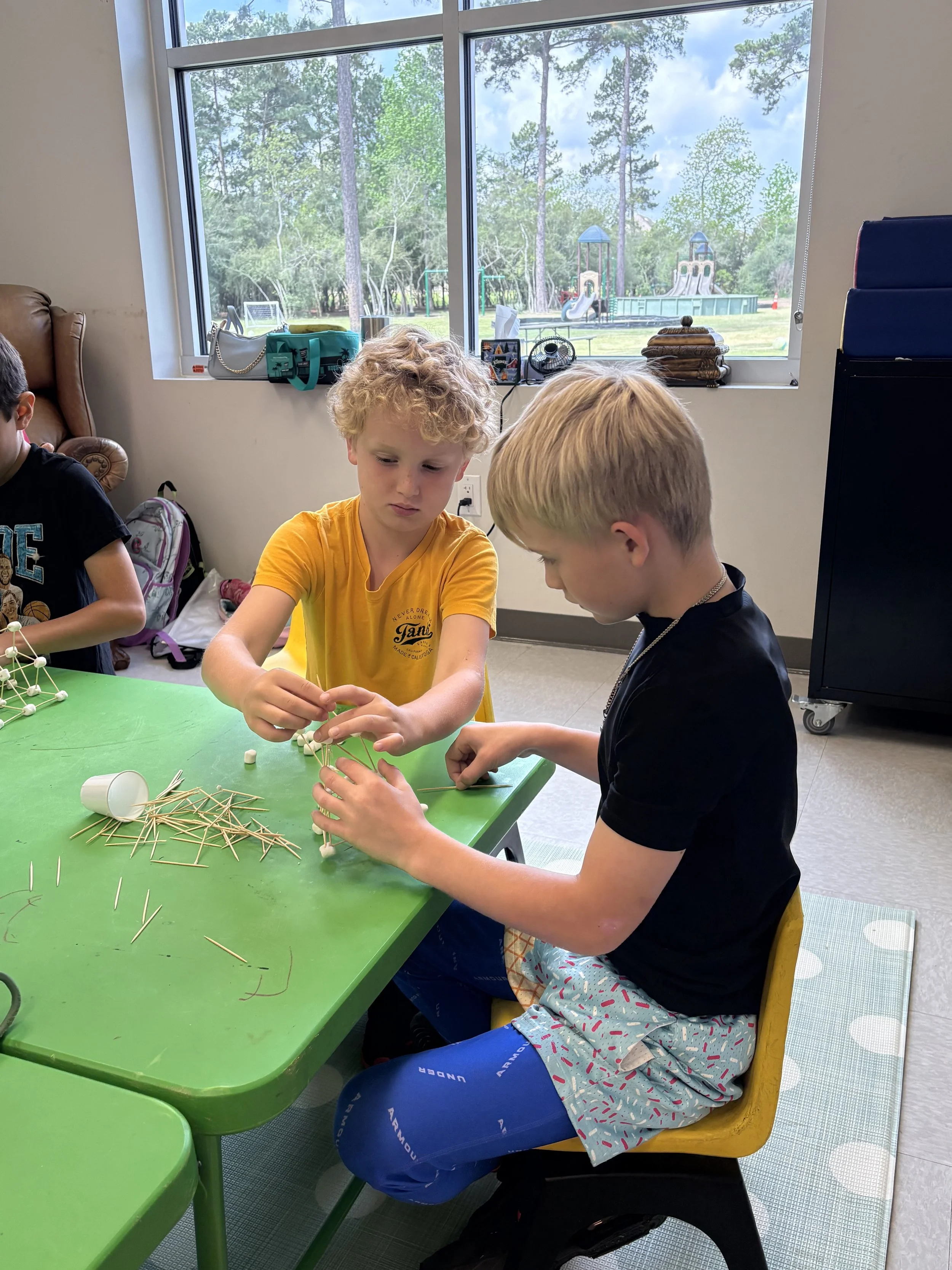 Two boys sitting at a green table, working on a craft project with toothpicks and marshmallows in a classroom near a window showing an outdoor playground with trees and slides.