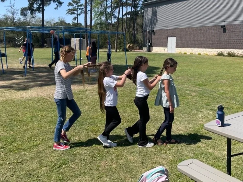 Five girls standing in a line outdoors, each holding the shoulders of the girl in front of her, creating a human train. They are on a grassy field with trees and a building in the background. In the foreground, there is a picnic table with a water bottle on it, and in the background, children are playing near a blue jungle gym.