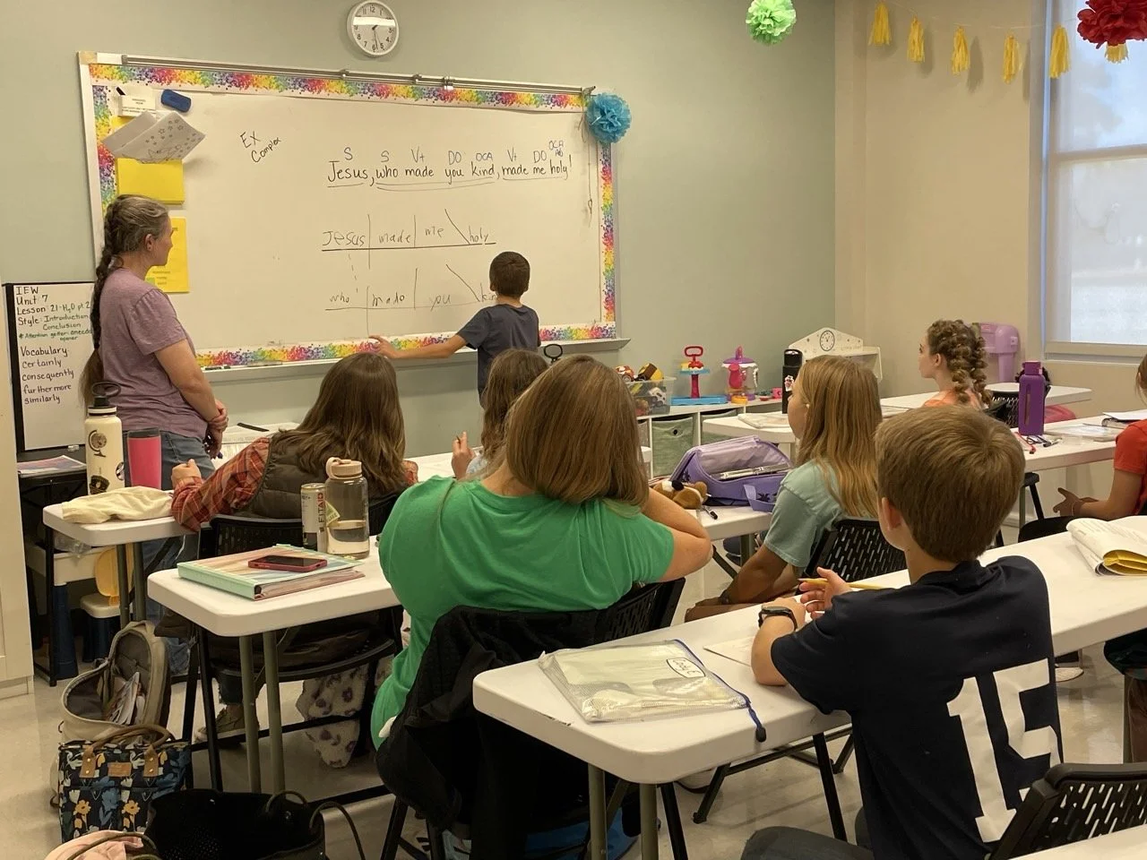 A classroom scene where a teacher and students are engaged in a lesson. The teacher stands near a whiteboard, which has a Bible verse written on it. Students are sitting at desks, facing the whiteboard, with notebooks and water bottles on their desks. Decorations hang from the ceiling, and there is a clock above the whiteboard showing the time.