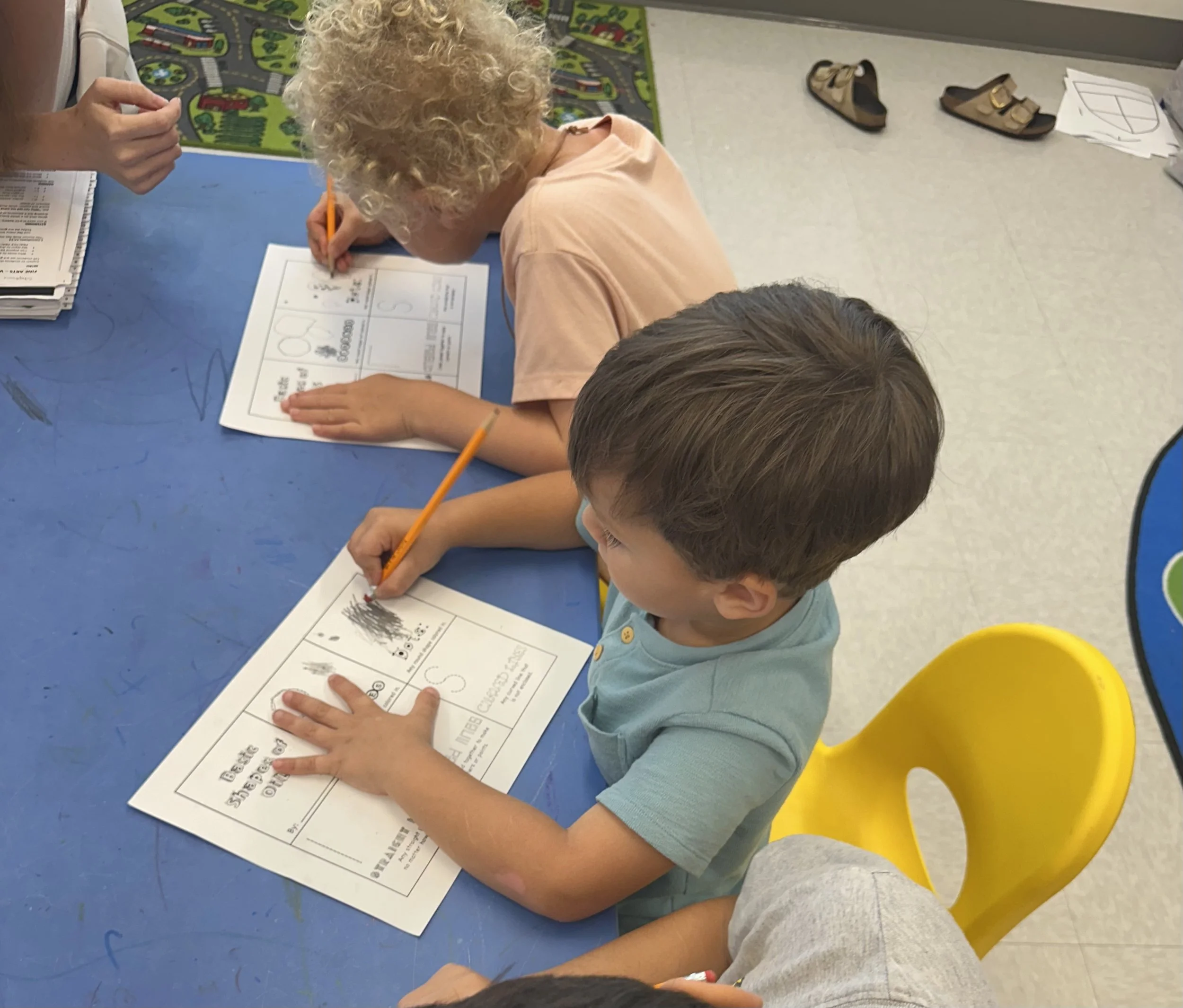 Two children sitting at a blue table, focusing on a worksheet, with one child using a pencil. There is a pair of sandals and papers on the floor nearby, and a colorful rug with a cityscape pattern in the background.