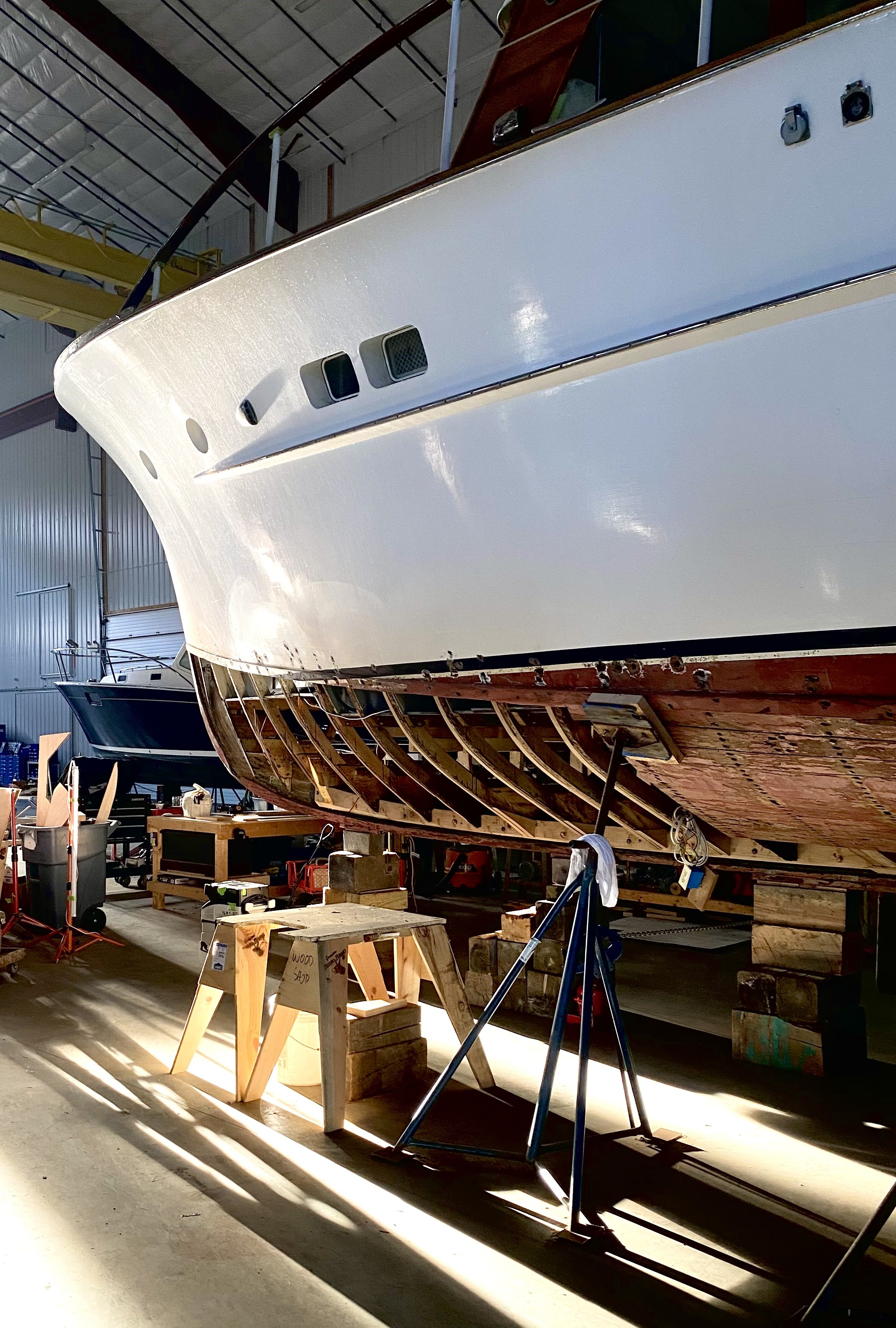 A large boat is under construction in a boat workshop. The boat is on supports and the hull is partially painted white with a visible wooden understructure. The workshop has tools, wooden blocks, and equipment around.