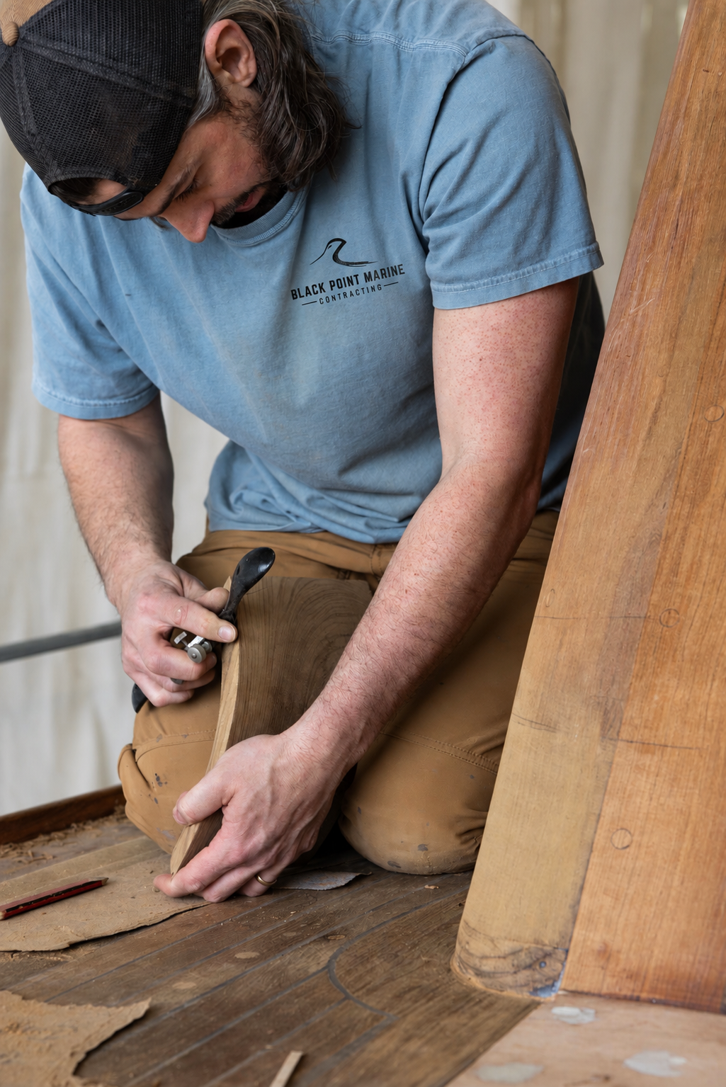A man wearing a blue t-shirt with 'Black Point Marine Contracting' logo and a black cap is kneeling and using a hand plane on a piece of wood during a woodworking project.