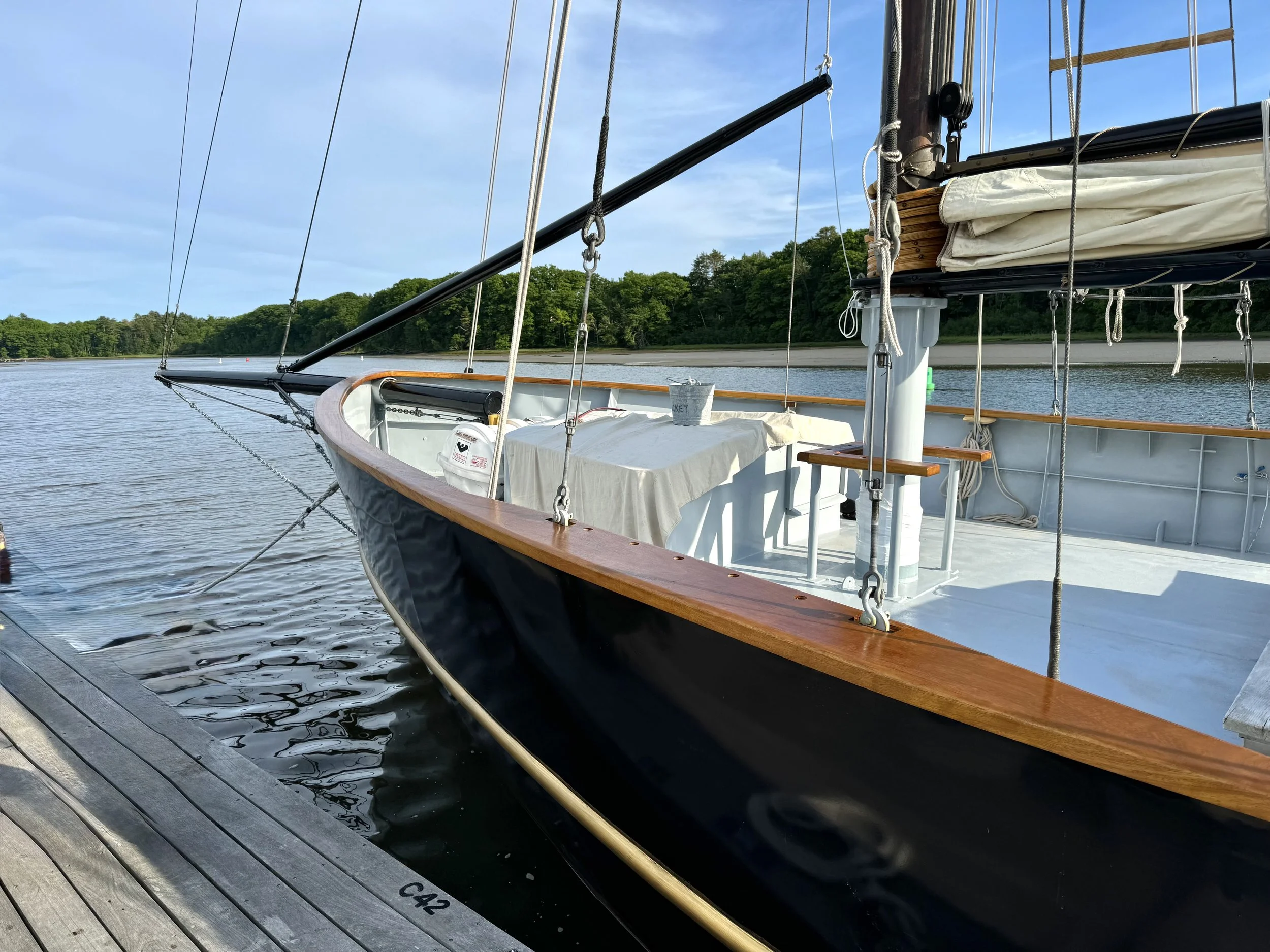 View of a sailboat moored at a dock on calm water with a background of green trees and a partly cloudy sky.