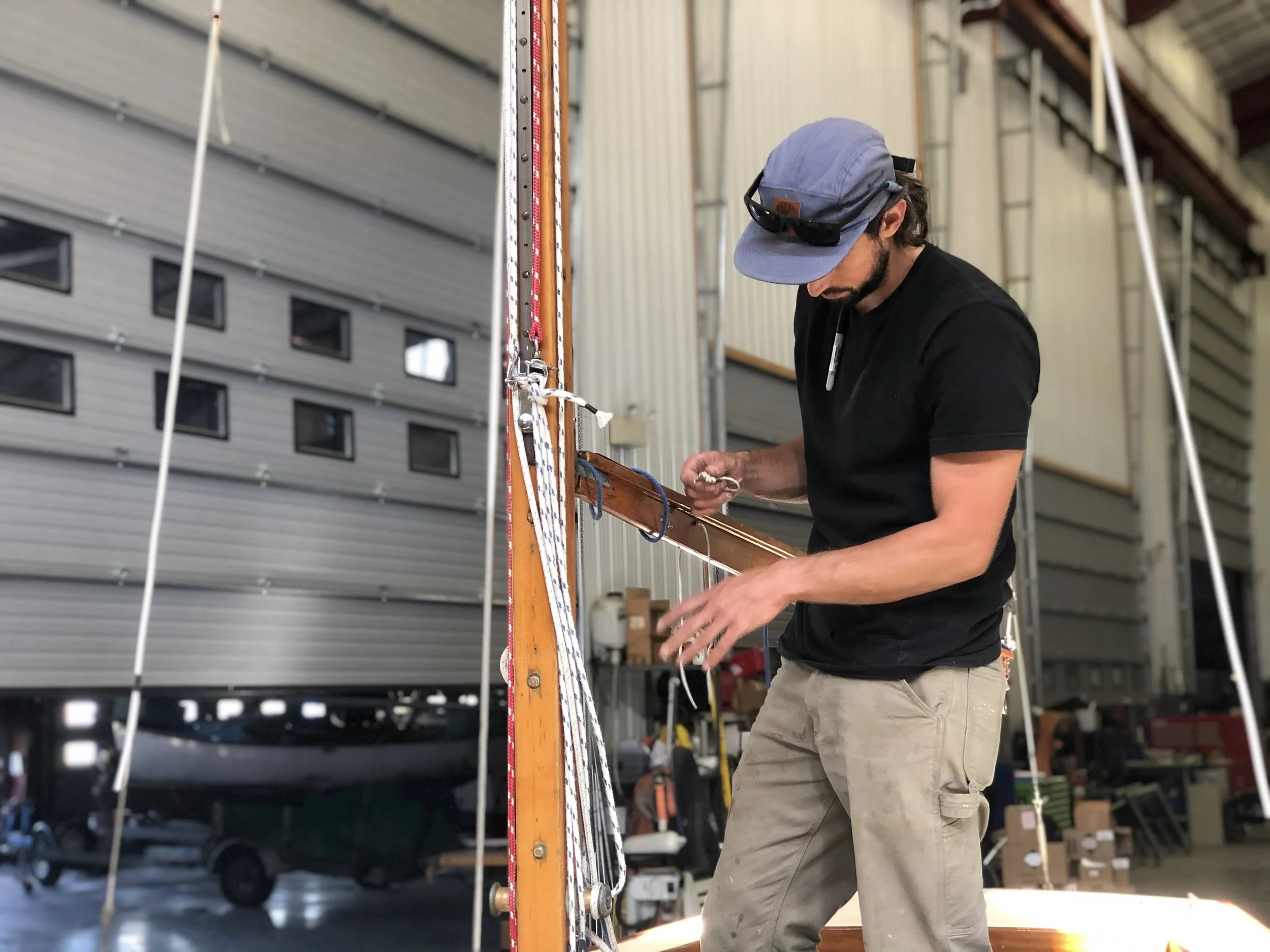 A man wearing a blue cap, sunglasses, and black shirt working in a warehouse with boats and storage shelves in the background.