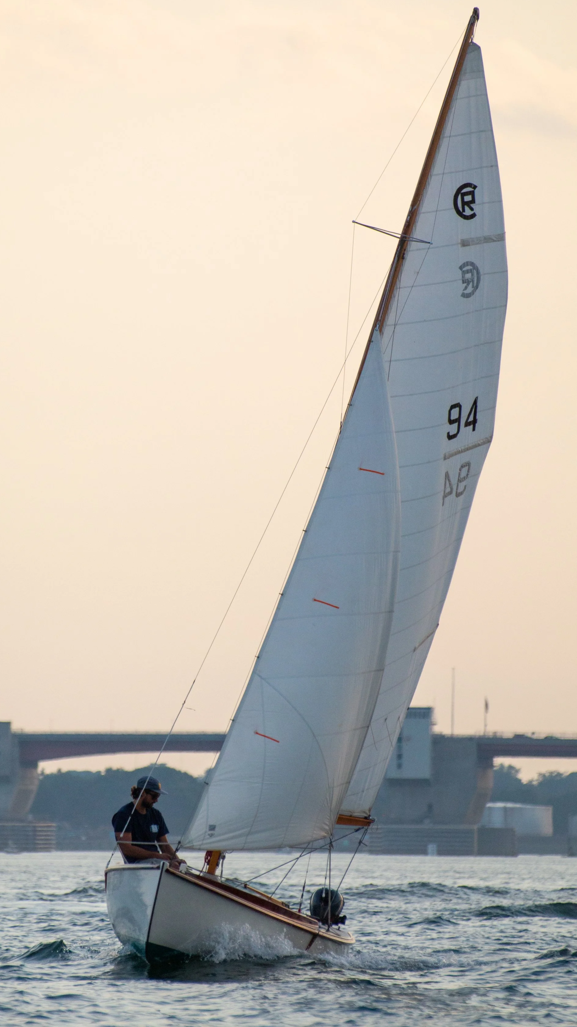 A sailboat with large white sails sailing on the water during sunset, with a bridge and city skyline in the background.