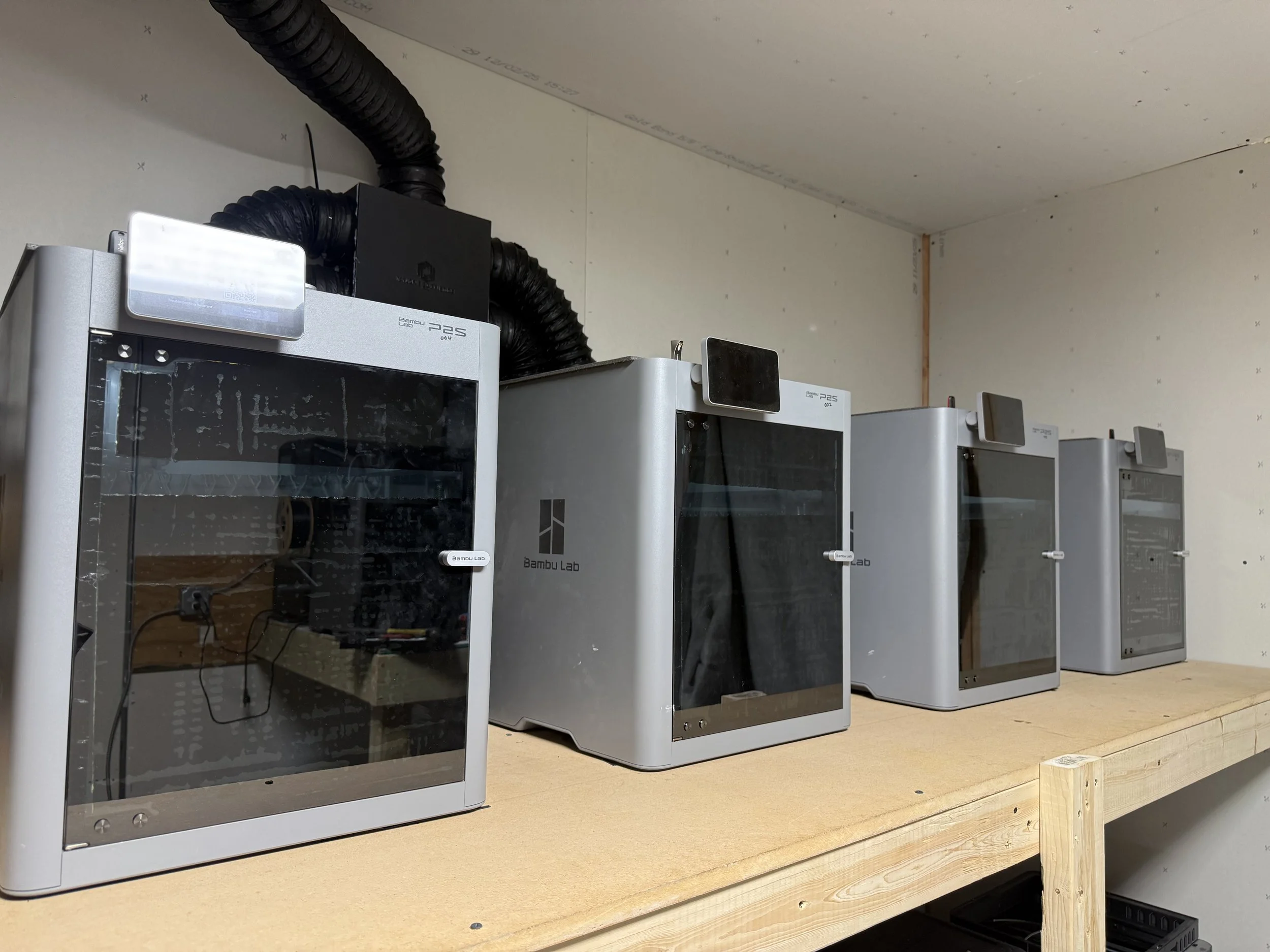 Three silver 3D printers labeled 'Bambu Lab' placed on a light wooden workbench in a room with plain white walls.