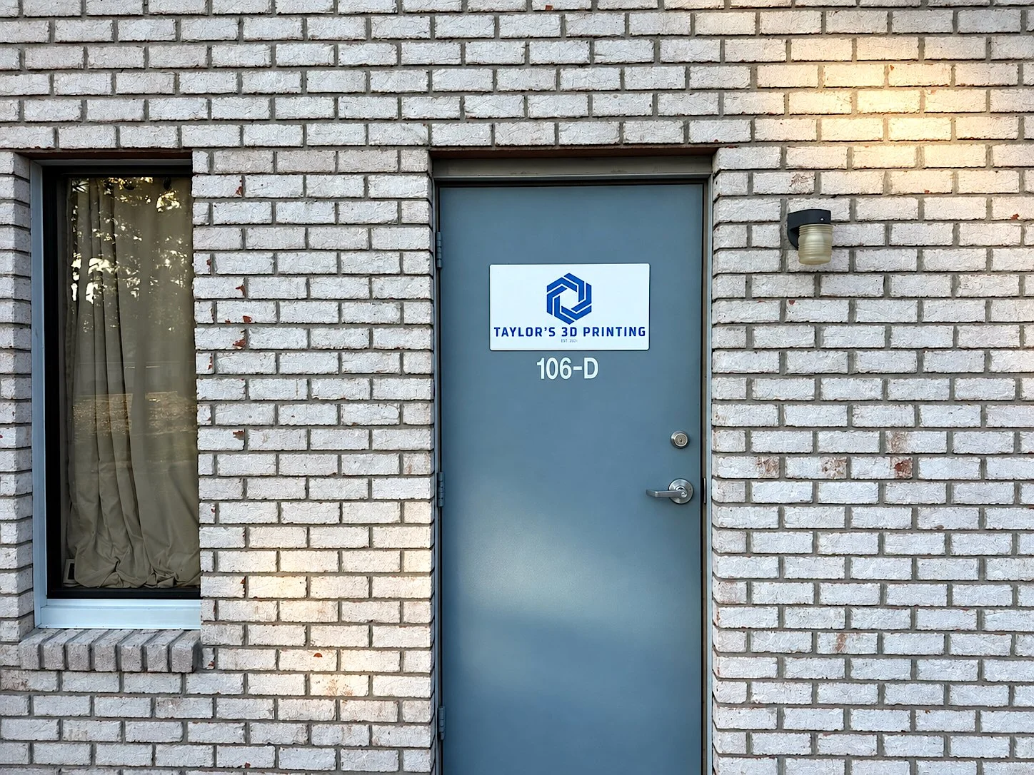Brick building exterior with a gray door and a sign for Taylor's 3D Printing, location 106-D, and a window with beige curtains to the left. An exterior light fixture is mounted on the wall to the right of the door.