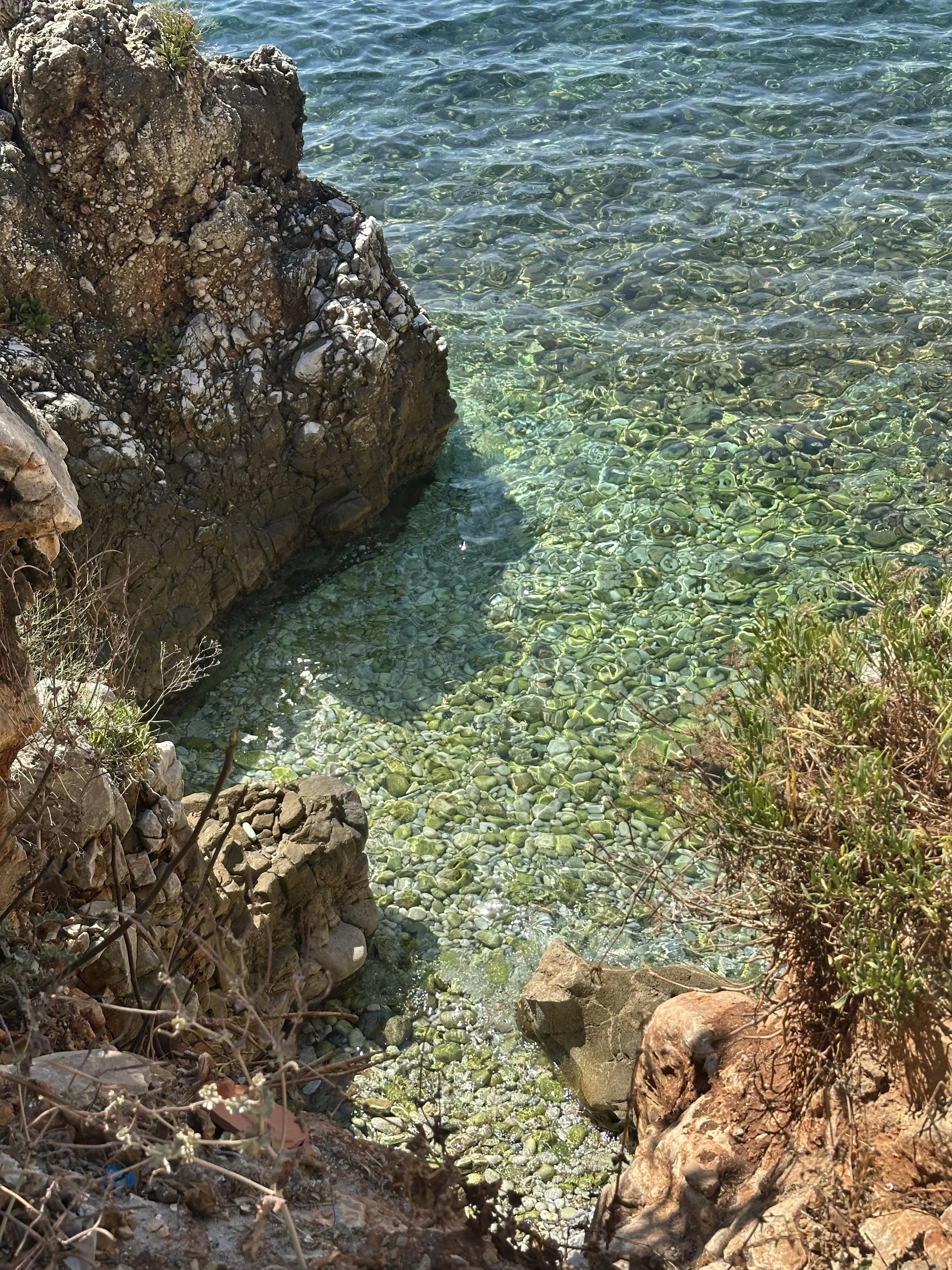 A rocky shoreline with clear water. Large rocks and smaller pebbles are visible along the coast, with some green plants near the rocks and the water is turquoise with gentle ripples.