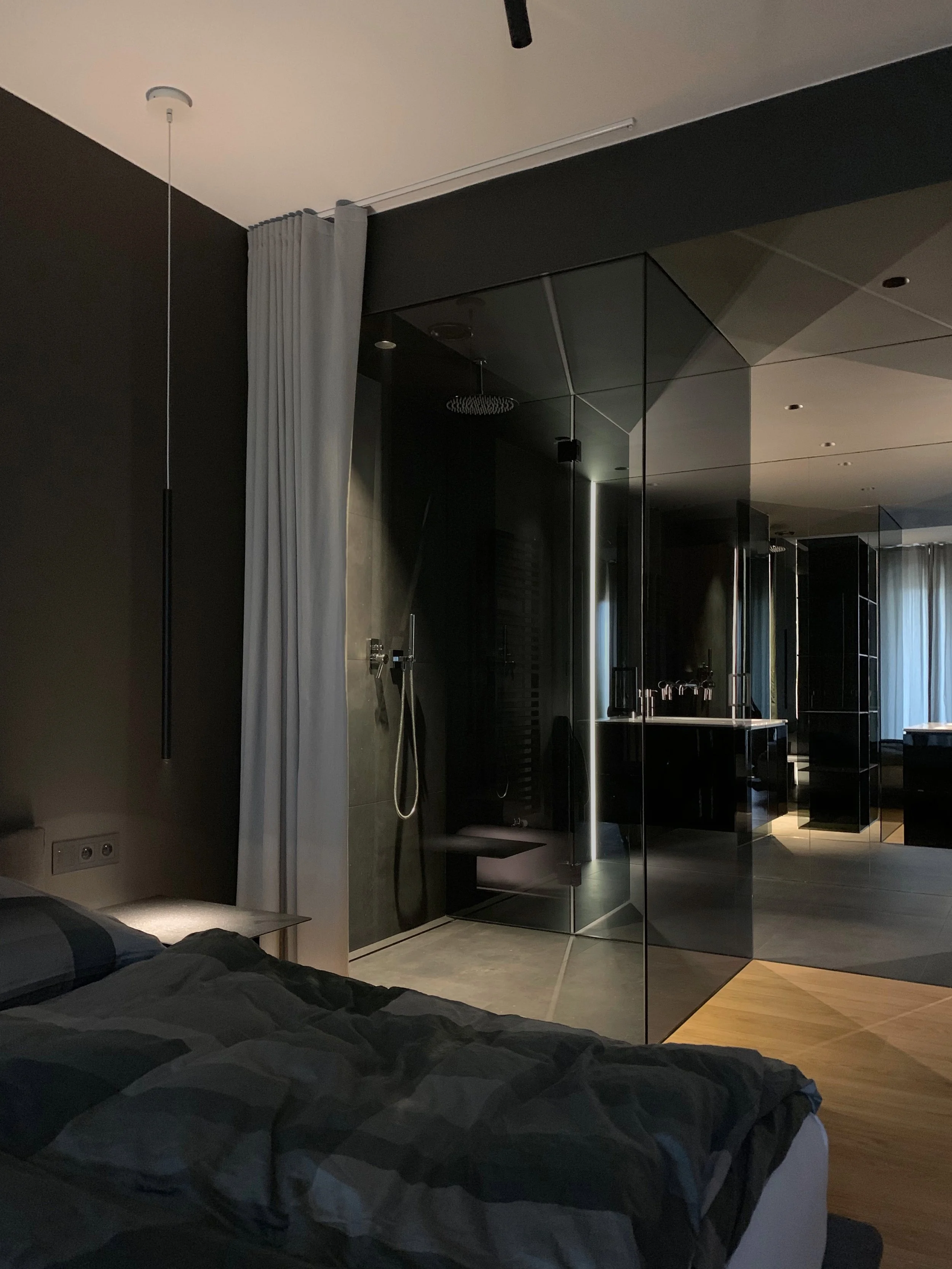 Modern bedroom with dark walls, black and gray bedding, a white curtain, and a glass-encased shower area visible through a glass wall.