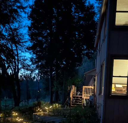 Nighttime scene of a house with illuminated windows and string lights in the yard, surrounded by trees.